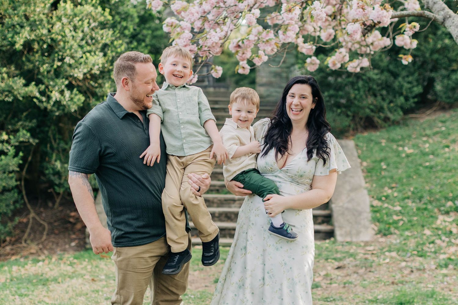 Happy family with two toddlers laughing under pink cherry blossoms in a lush green garden.