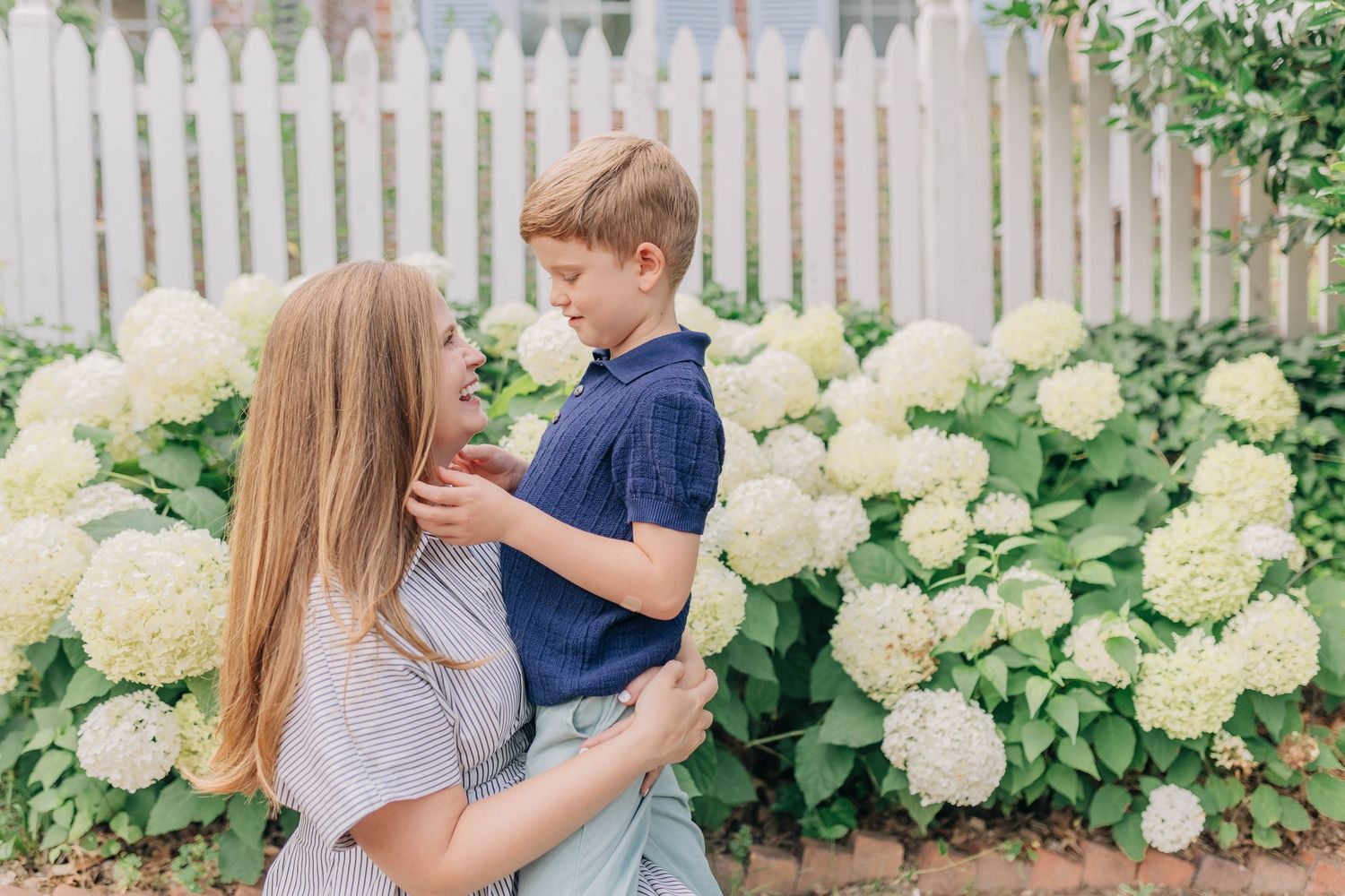 A mother holds her young son among white hydrangea bushes near a white picket fence outdoors.
