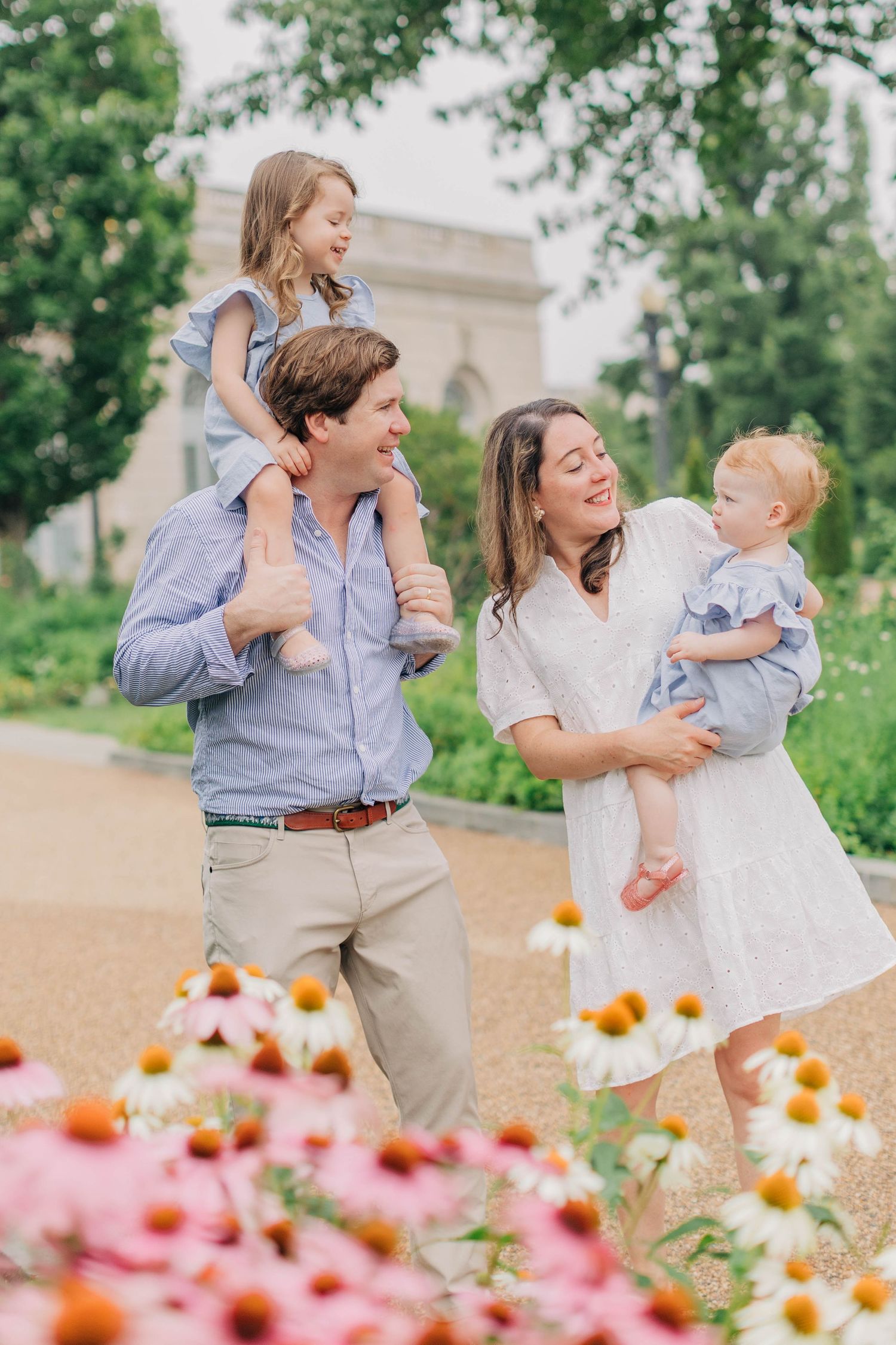 A happy family with two young children poses among blooming flowers in a lush garden park setting.