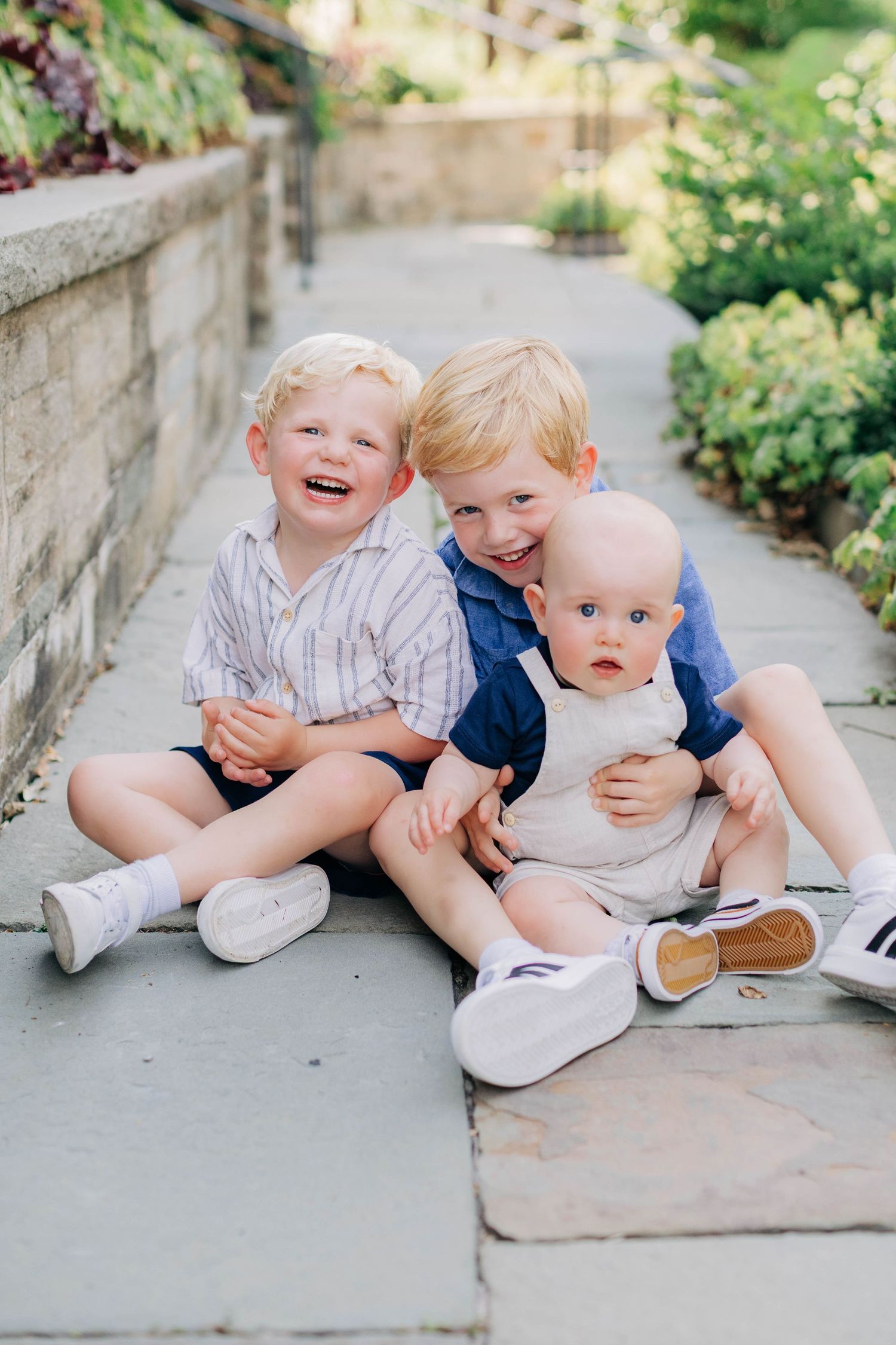 Three young blonde boys sitting together on a stone path in a garden, smiling and hugging.