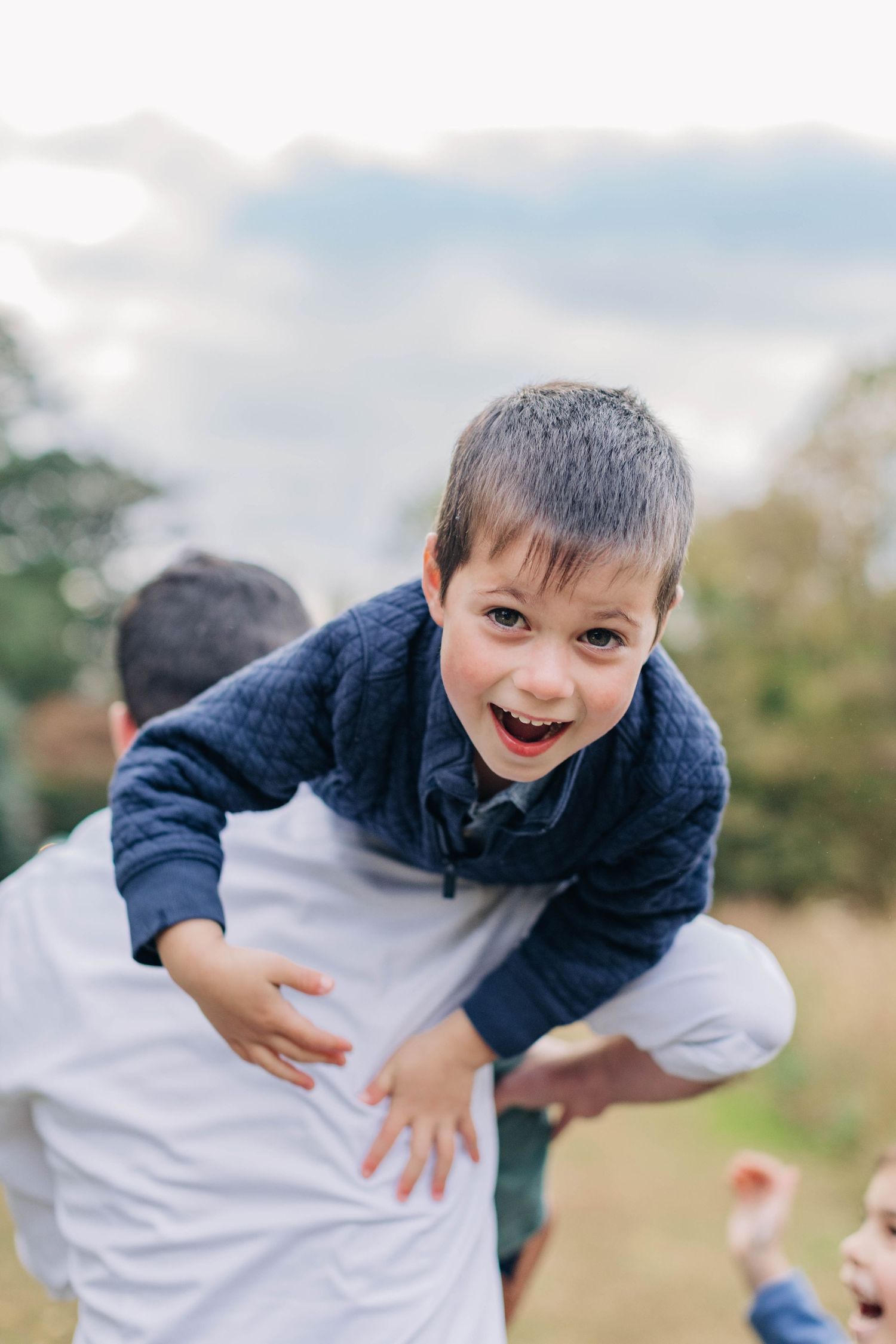 A joyful young boy in a blue sweater laughs while being lifted outdoors with a cloudy sky backdrop.