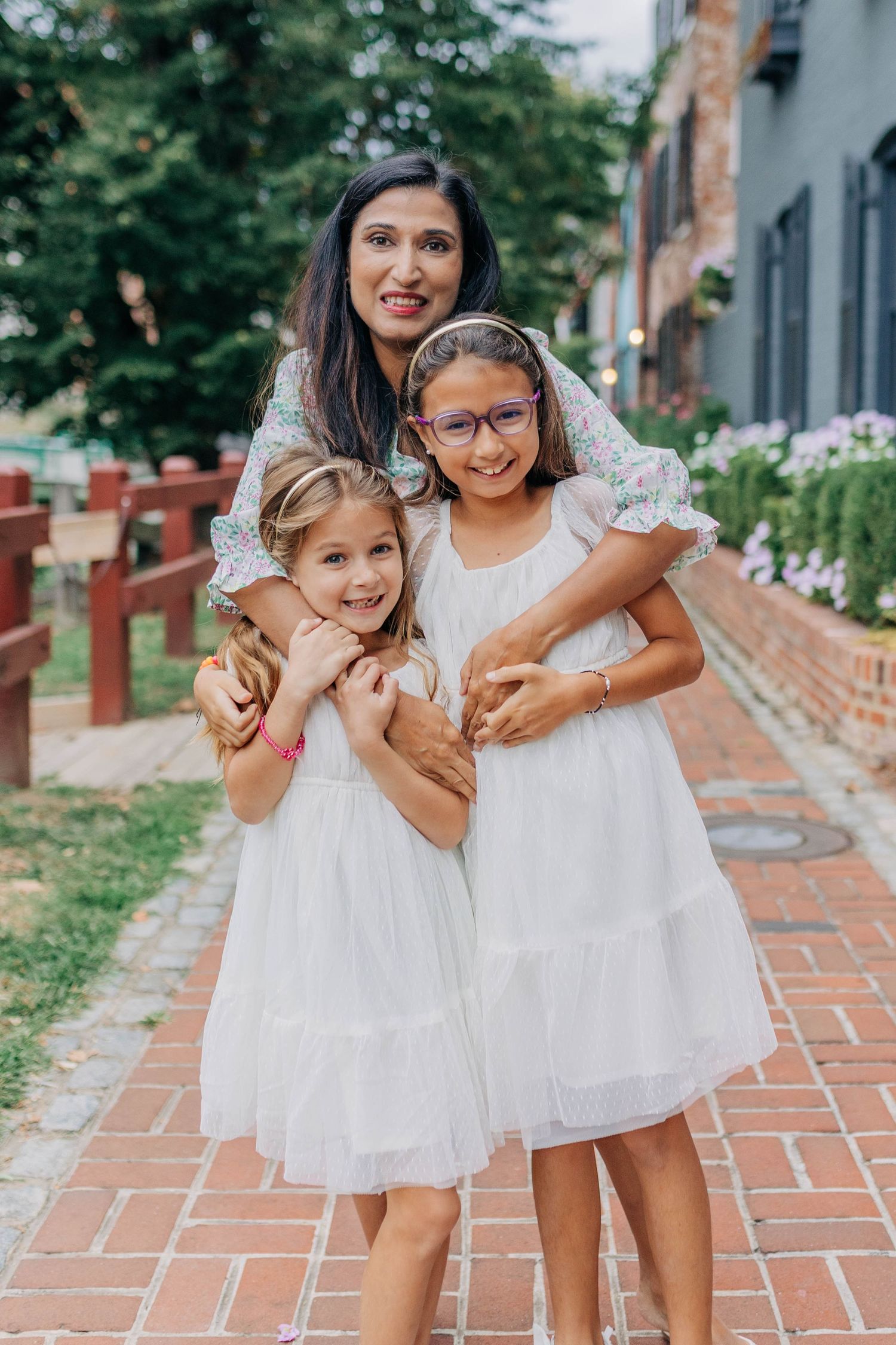 A woman in a floral dress hugs two girls in white dresses on a brick path outdoors.