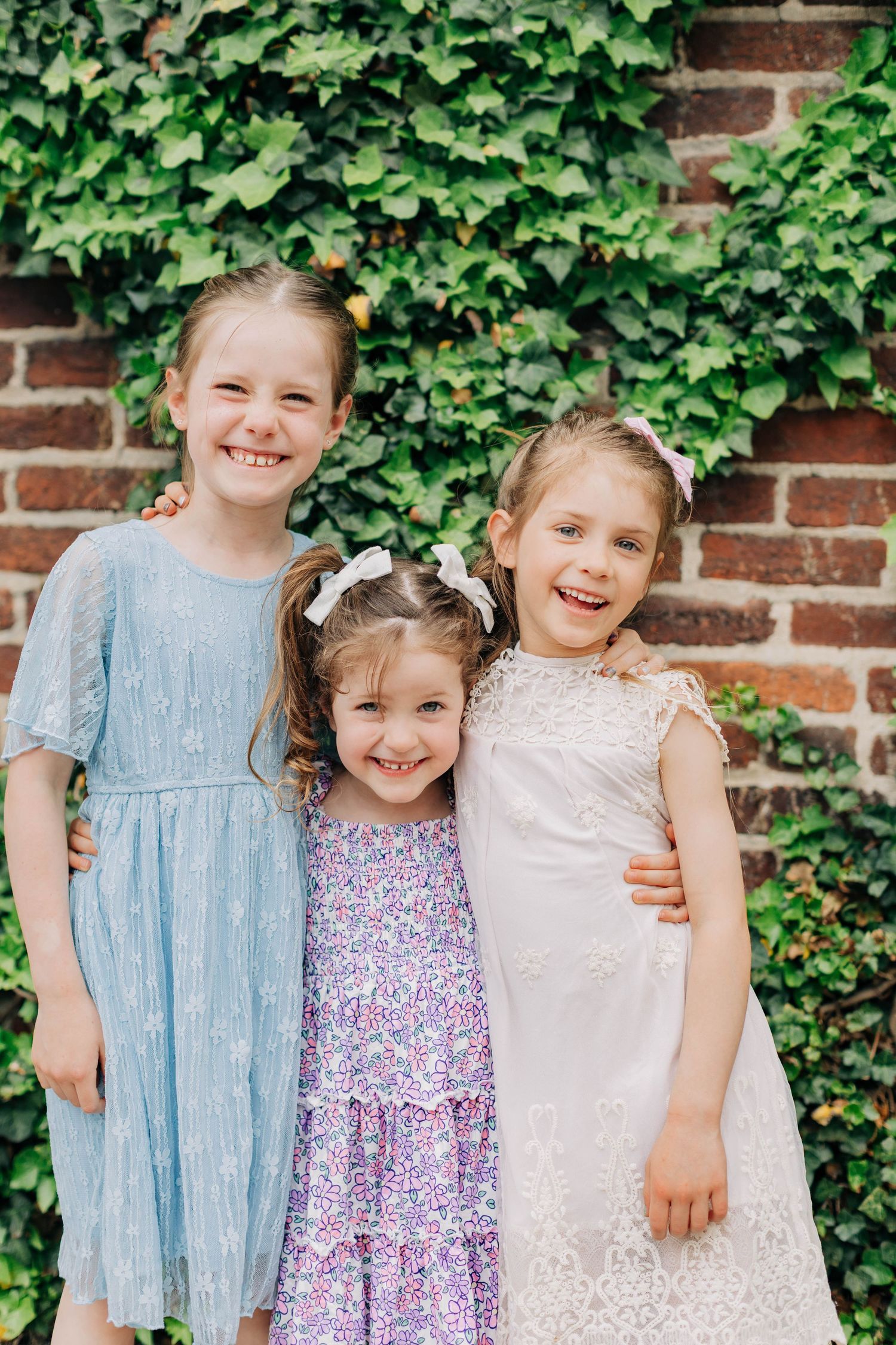 Three smiling girls in pastel dresses pose together in front of a brick wall covered in green ivy.