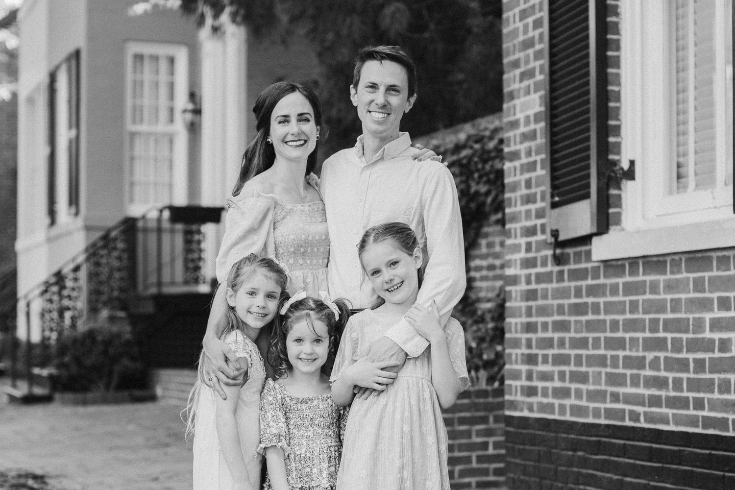 Black and white family portrait of parents with three young daughters smiling outdoors near a brick building.