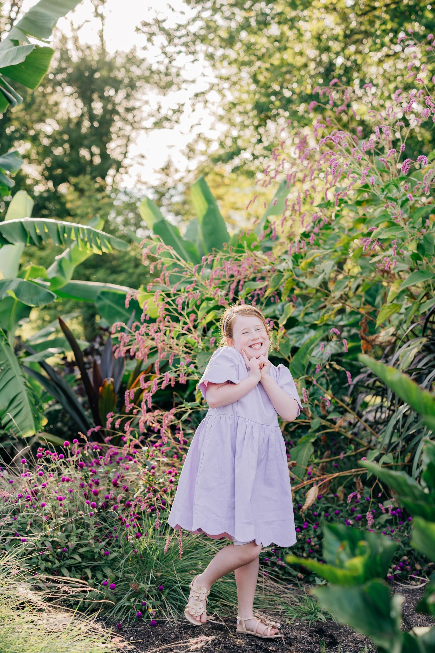 Young girl in lavender dress smiling joyfully among lush tropical garden flowers and greenery in warm sunlight.