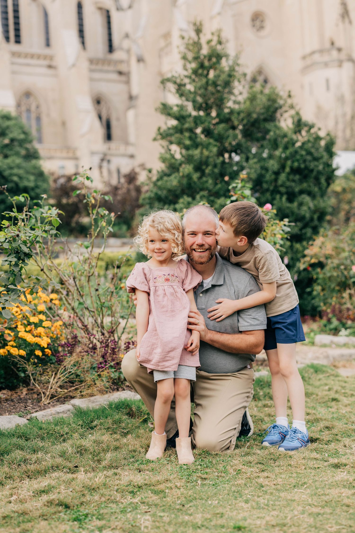 A smiling father kneels in a garden hugging his young daughter and son, with a stone cathedral in the background.