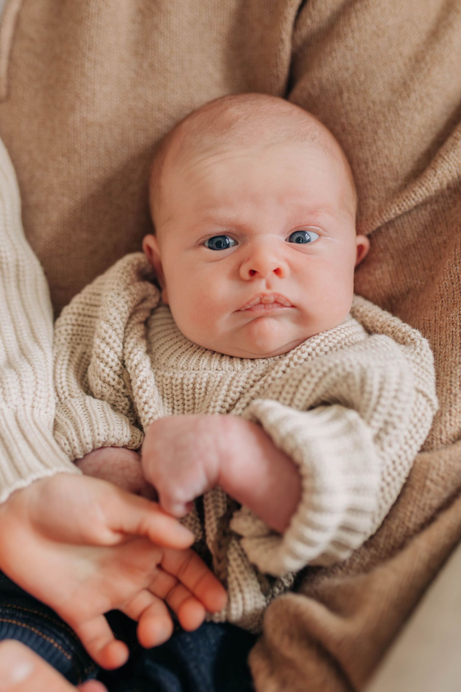 A baby wearing a cream knit sweater lies on soft brown fabric while being gently touched.