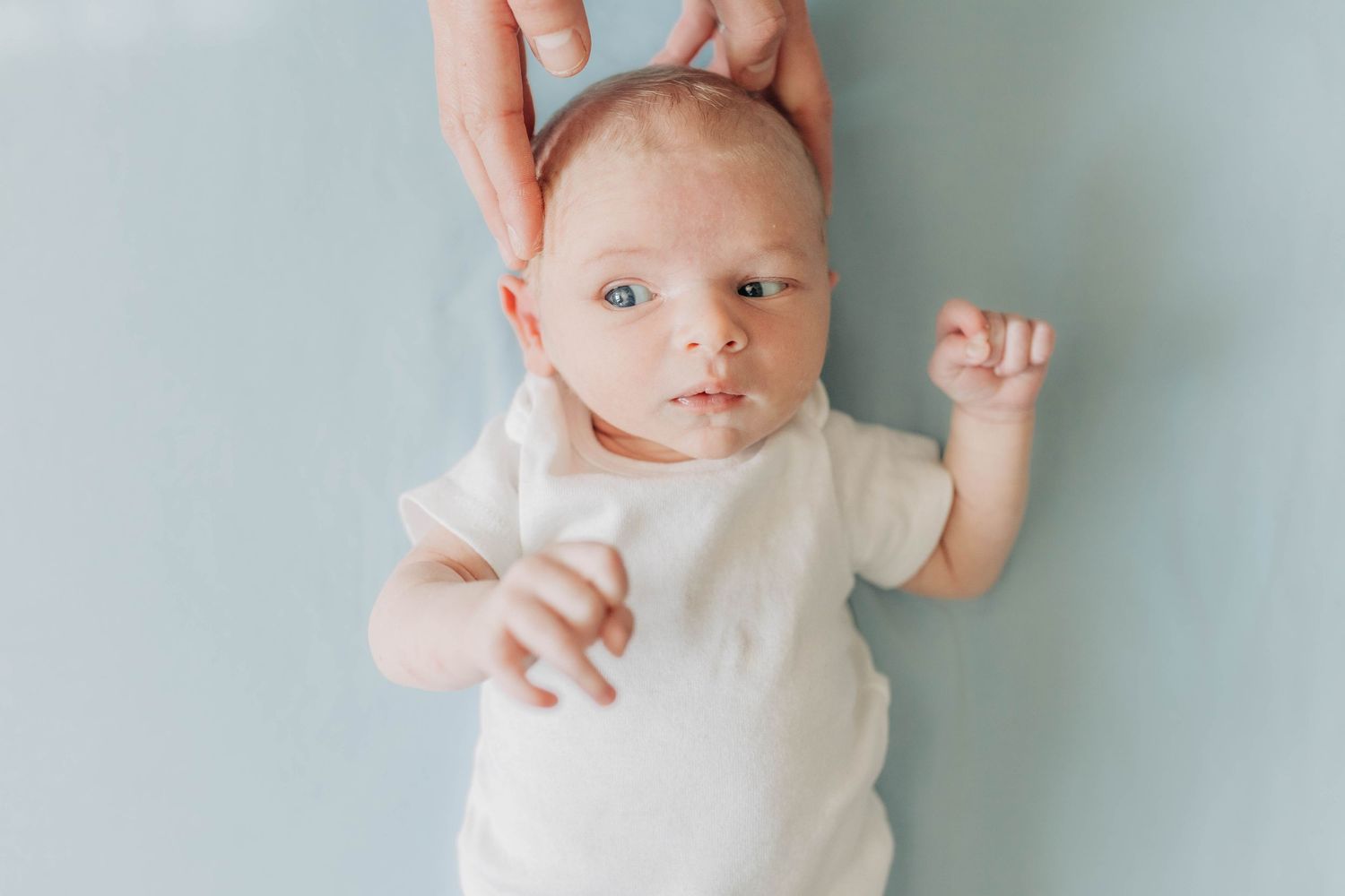 Baby in white onesie reaching up while being held against mint green wall.