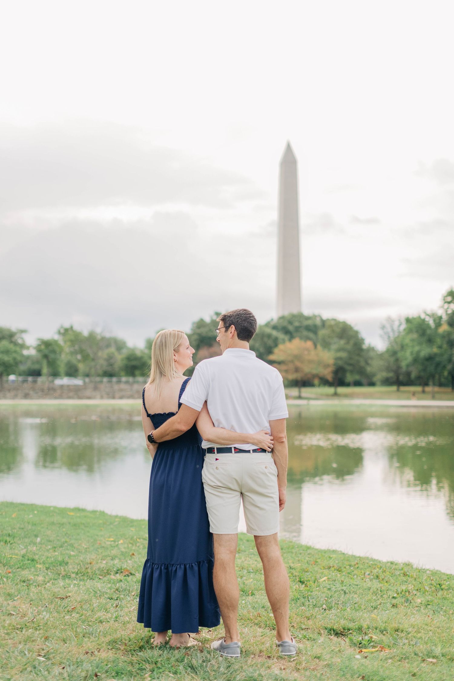 A couple stands together by a lake with the Washington Monument visible in the background.