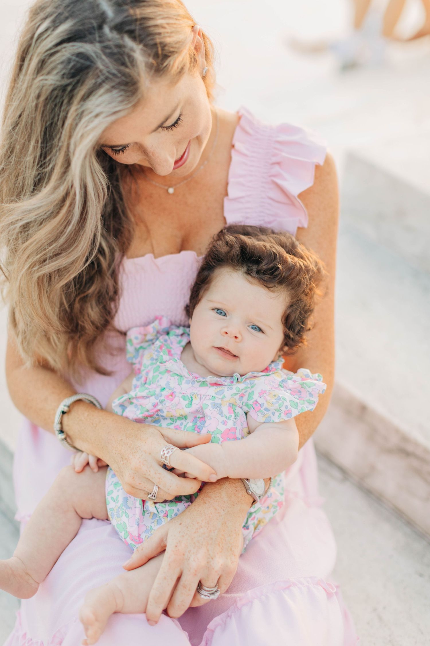 Parent in pink dress holding baby in floral outfit during outdoor photoshoot.