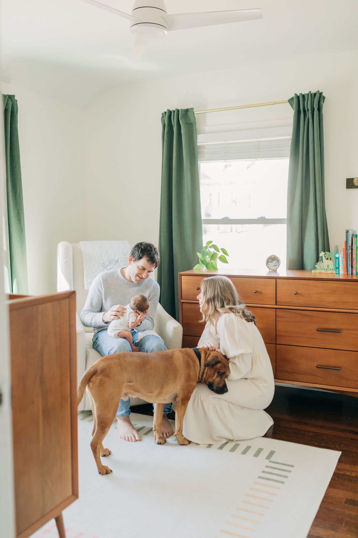 Family dog watches curiously as parent holds baby in bright nursery room.