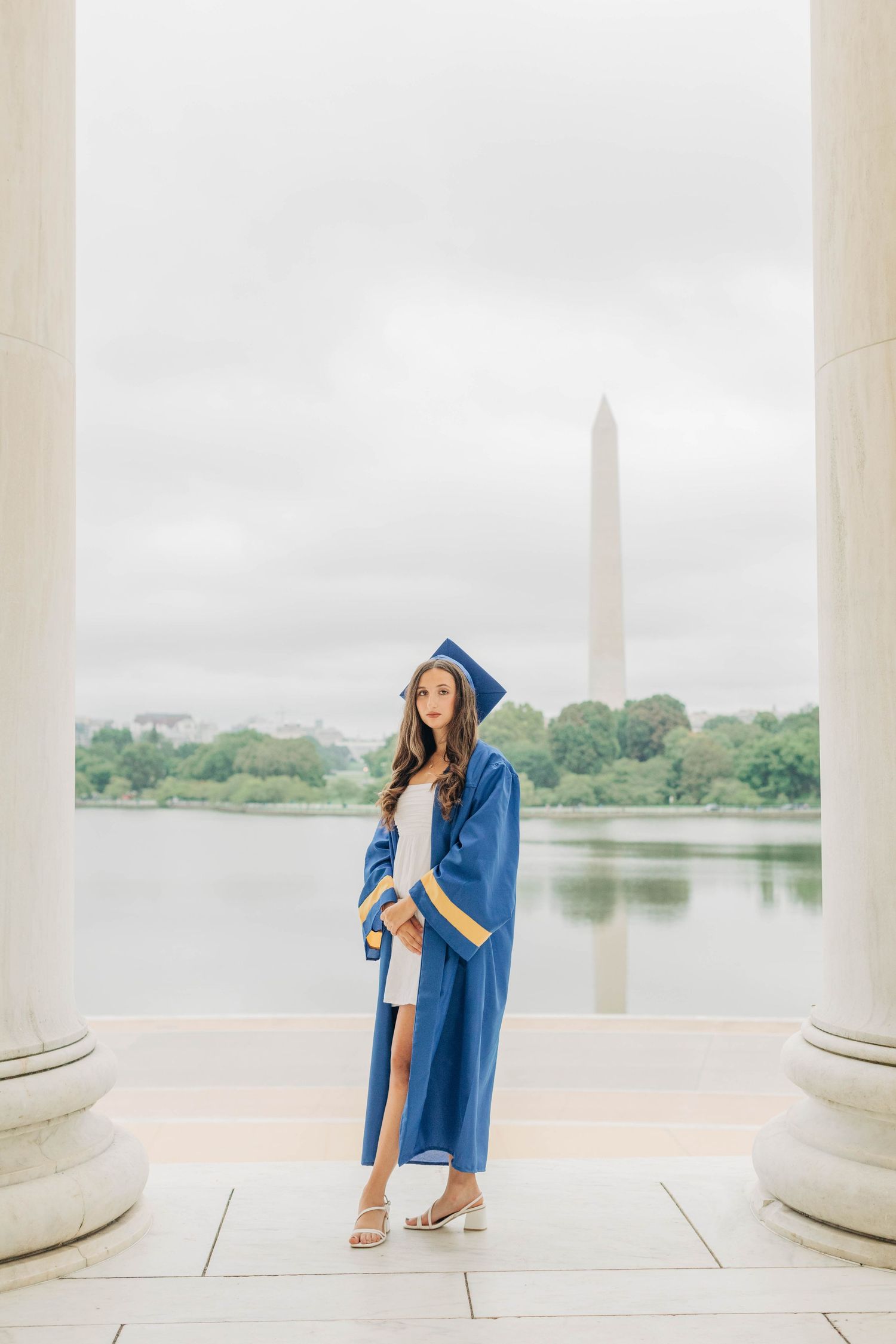 Graduate in blue cap and gown poses between columns with Washington Monument in background.