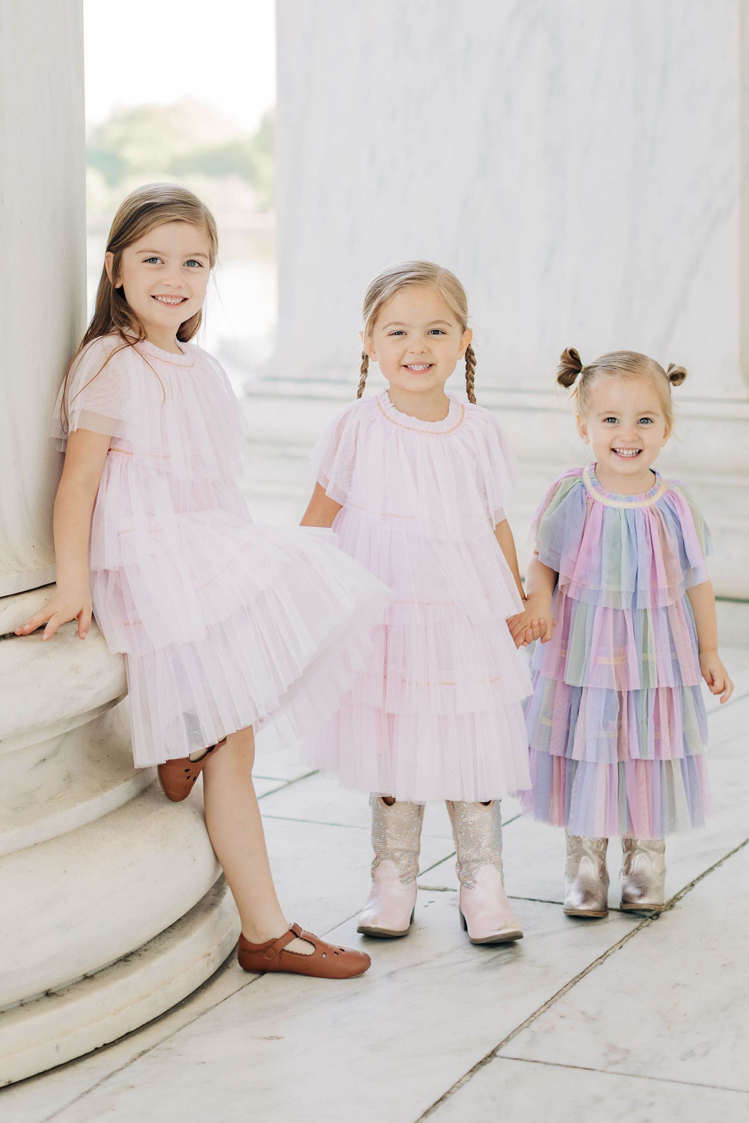 Three little girls in pink and pastel tiered tulle dresses pose together between marble columns.