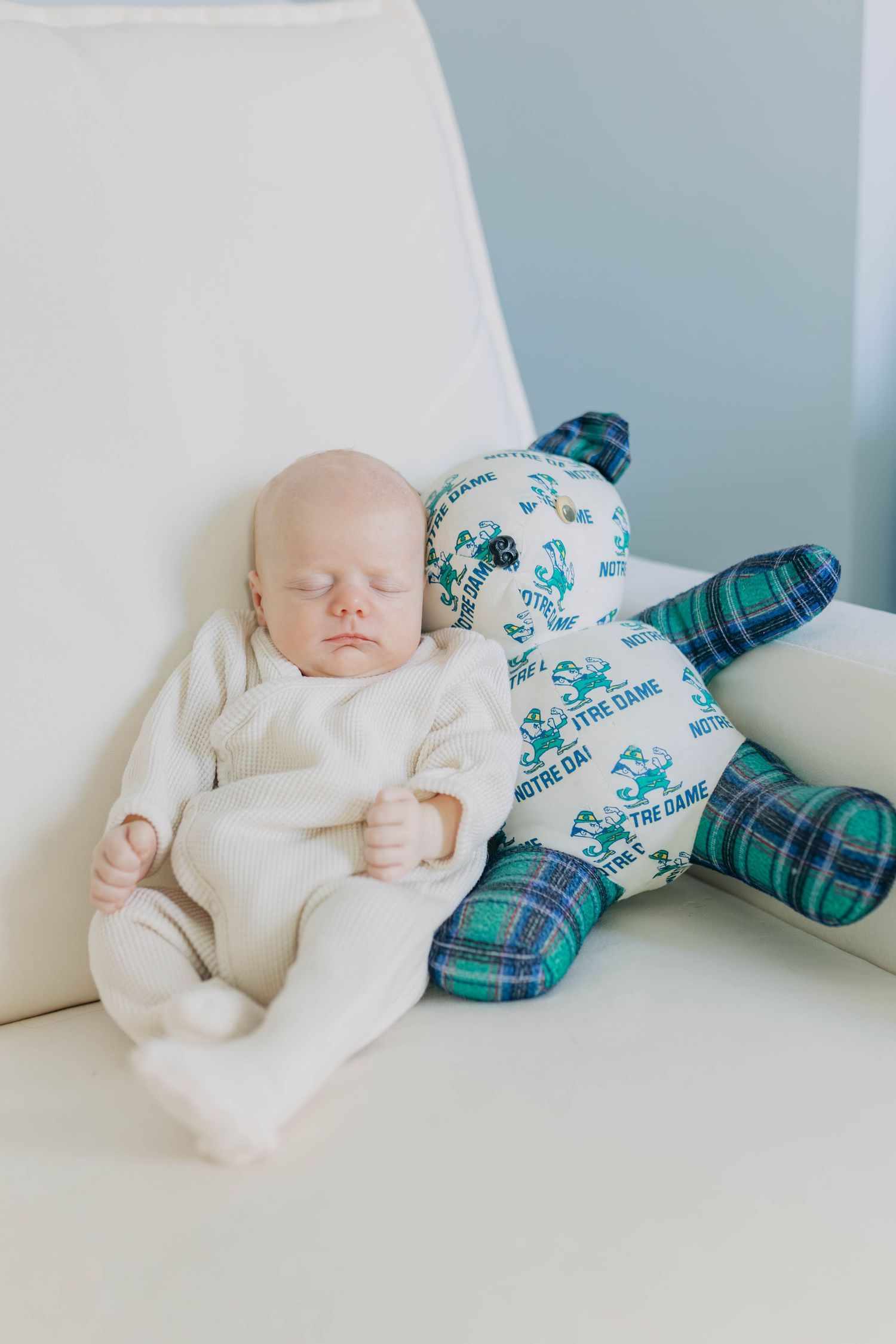 A sleeping newborn in white pajamas resting against a Notre Dame themed stuffed bear toy.
