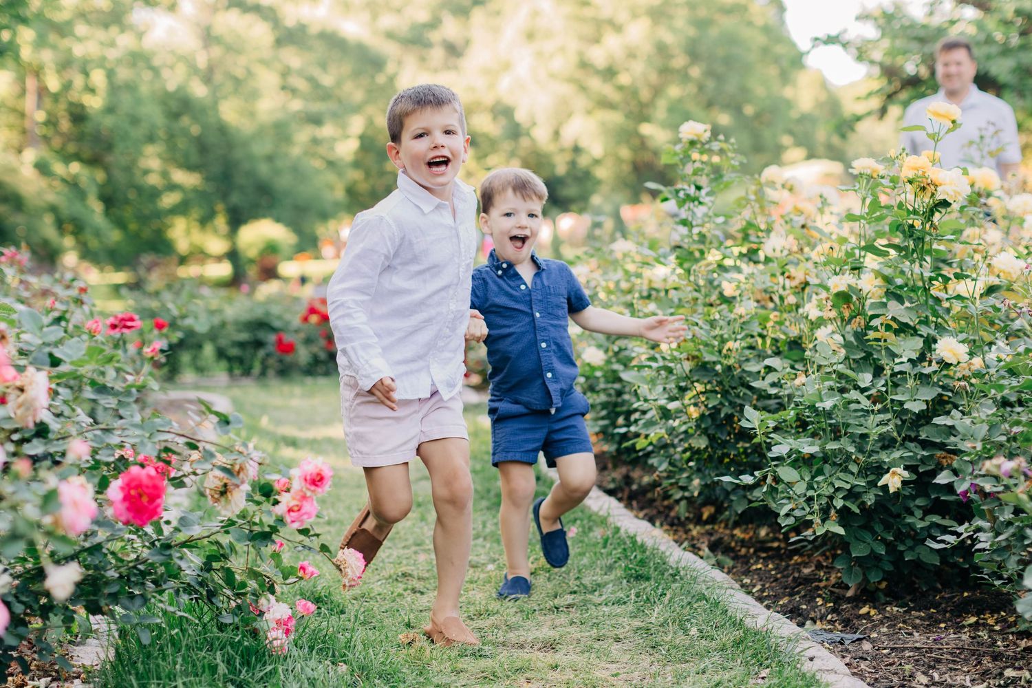Two laughing boys in white and blue outfits run joyfully through a vibrant rose garden.