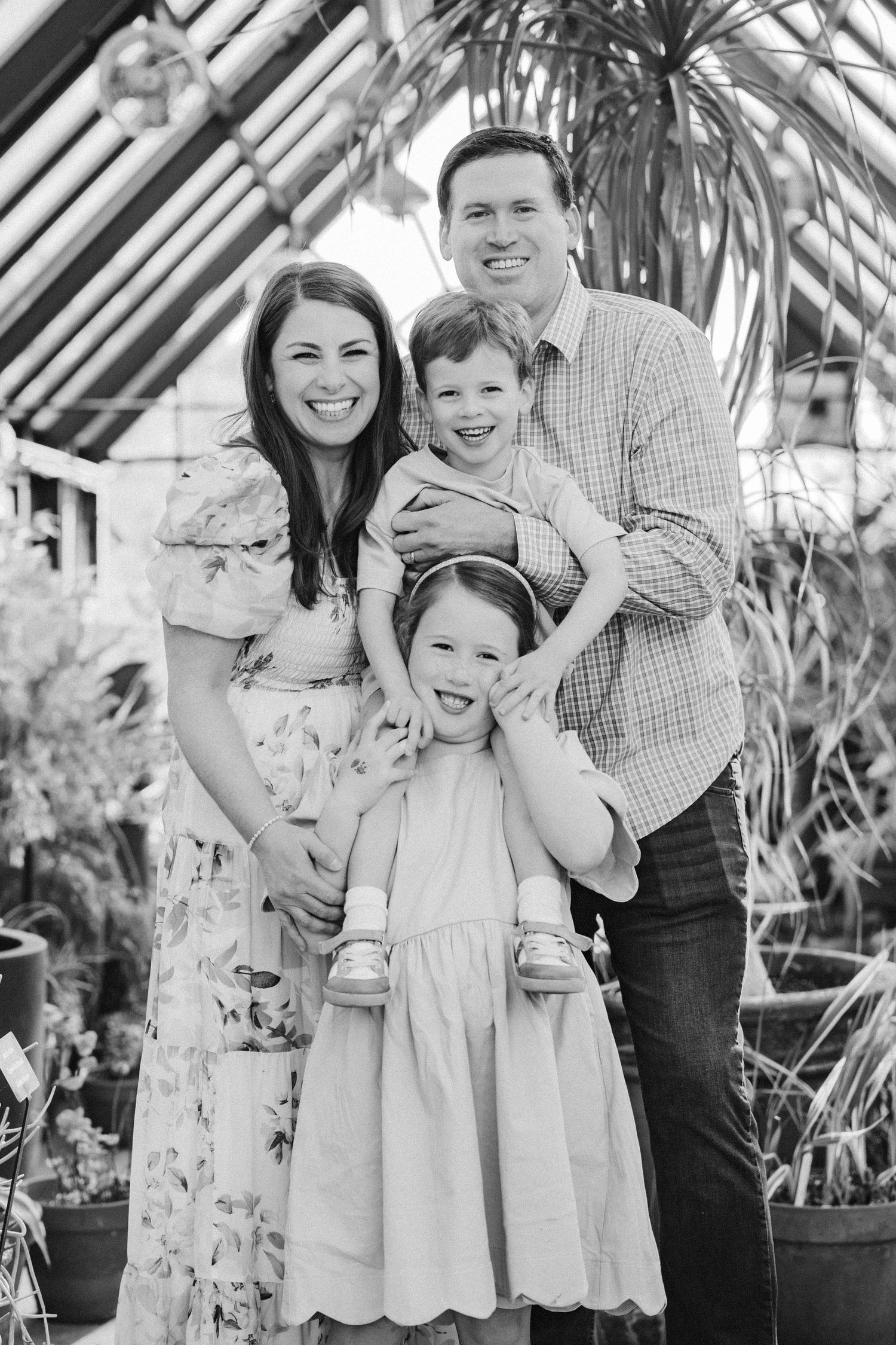 A black and white family portrait of parents and two children smiling in a greenhouse.