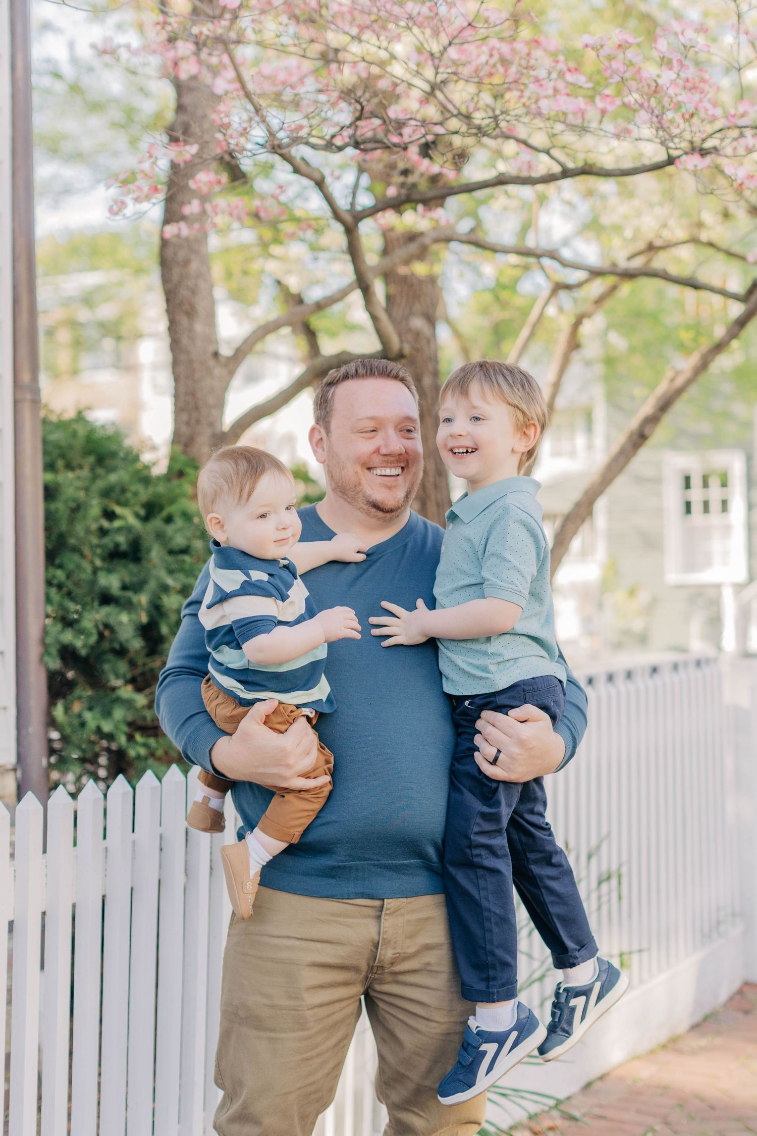 A smiling father holds two young boys in front of a white picket fence with blooming pink trees in spring.