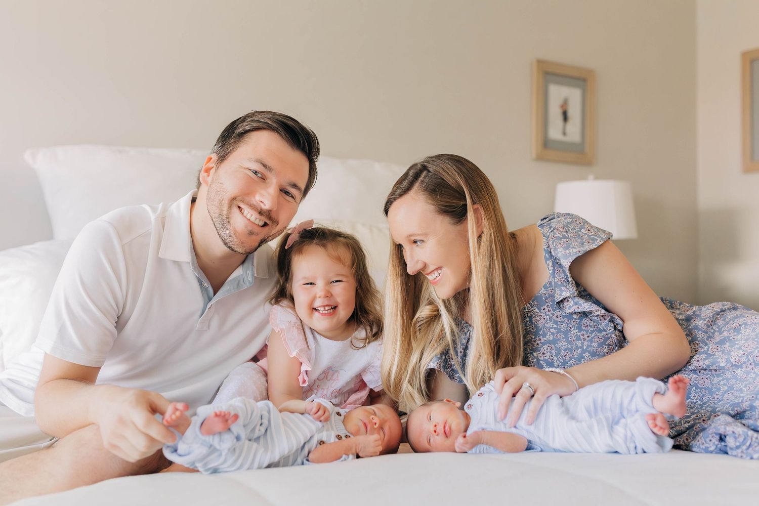 Happy family with parents, toddler girl, and newborn twins lying together on a white bed smiling.