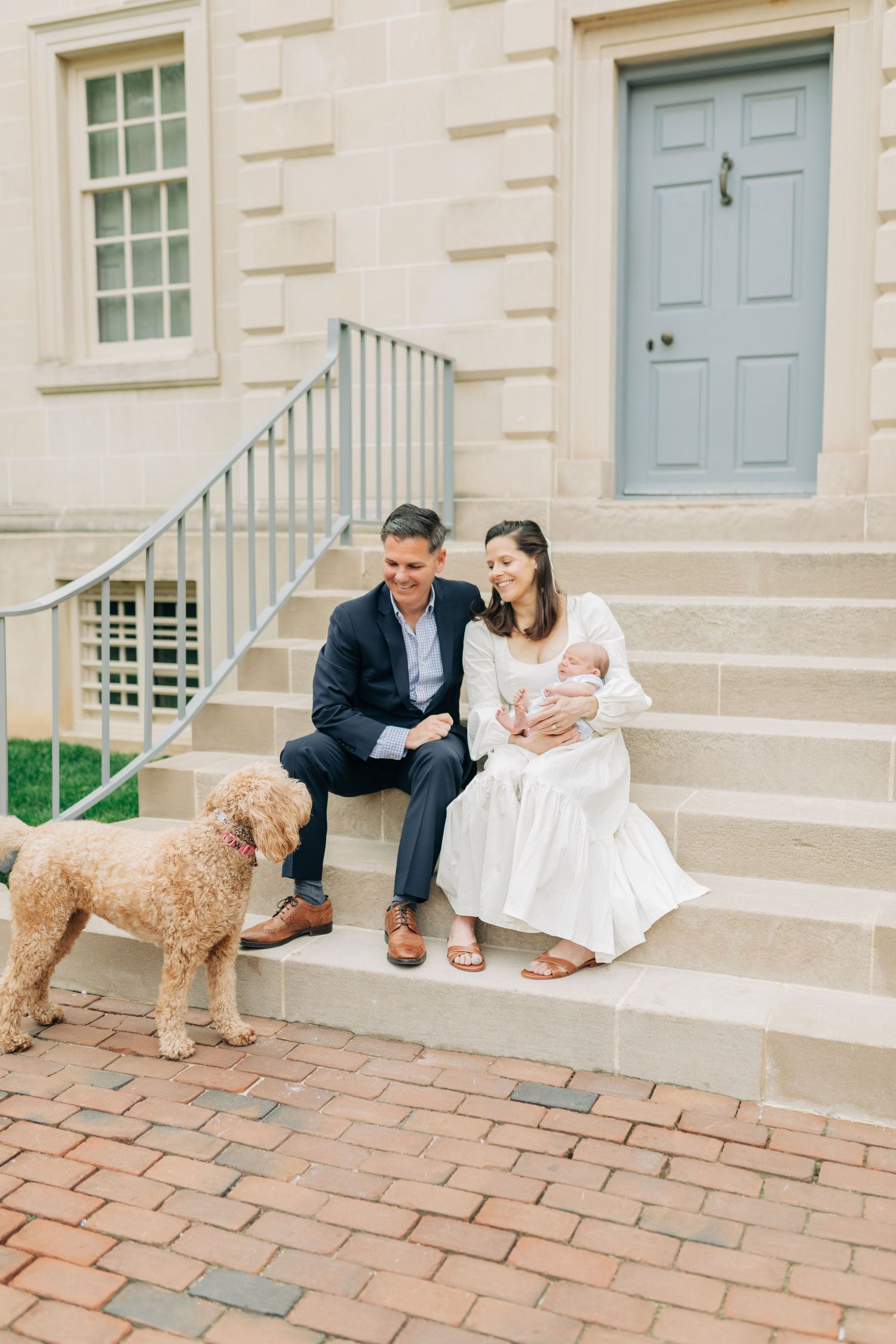 A couple and baby sit on stone steps with their fluffy dog beside a blue door.