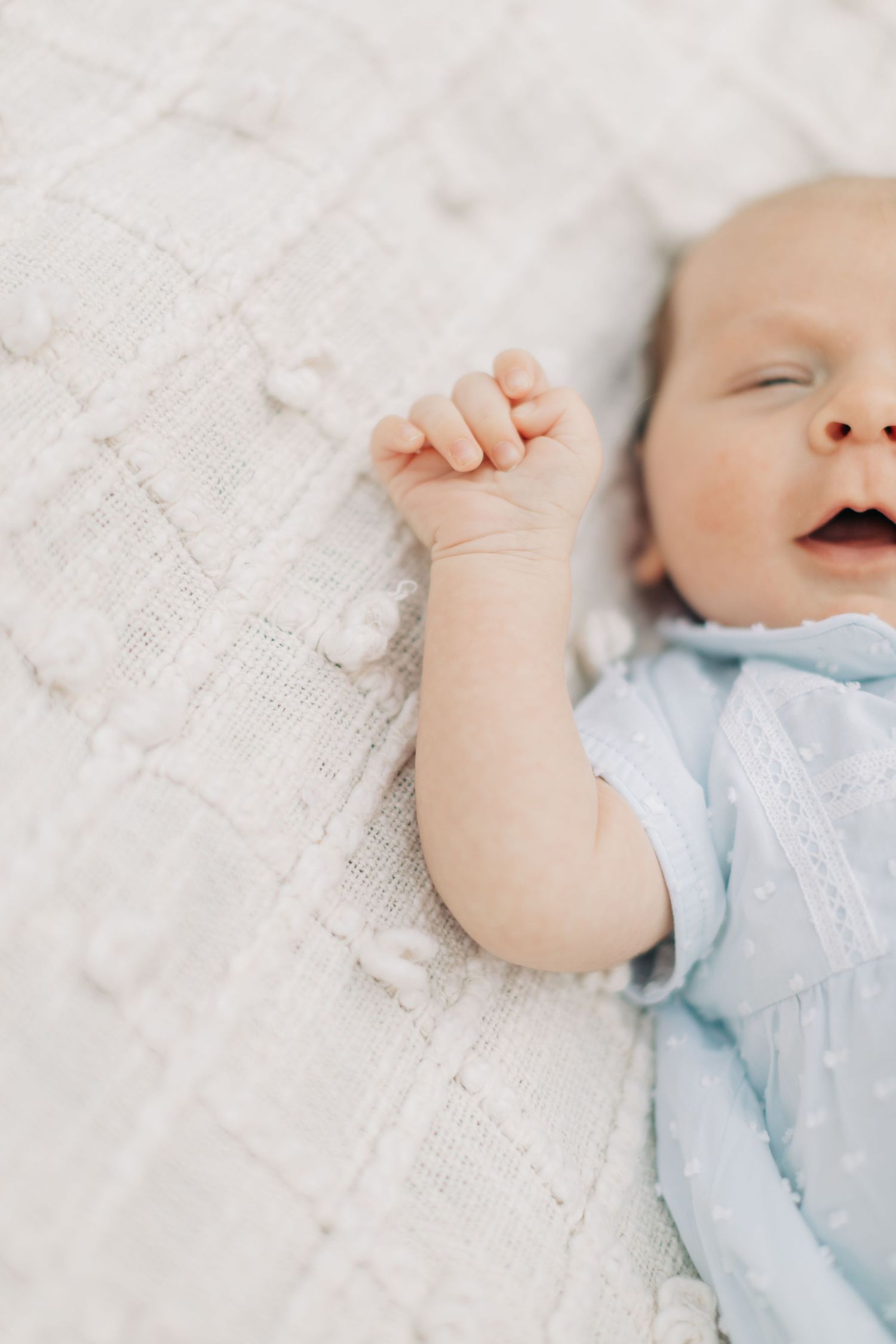 Baby in blue outfit with raised arm lying on white blanket.