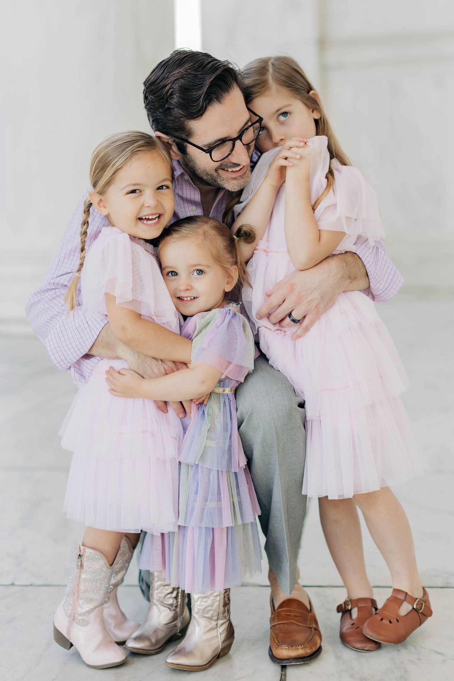 A father in glasses lovingly hugs his three young daughters in matching pink tulle dresses indoors.