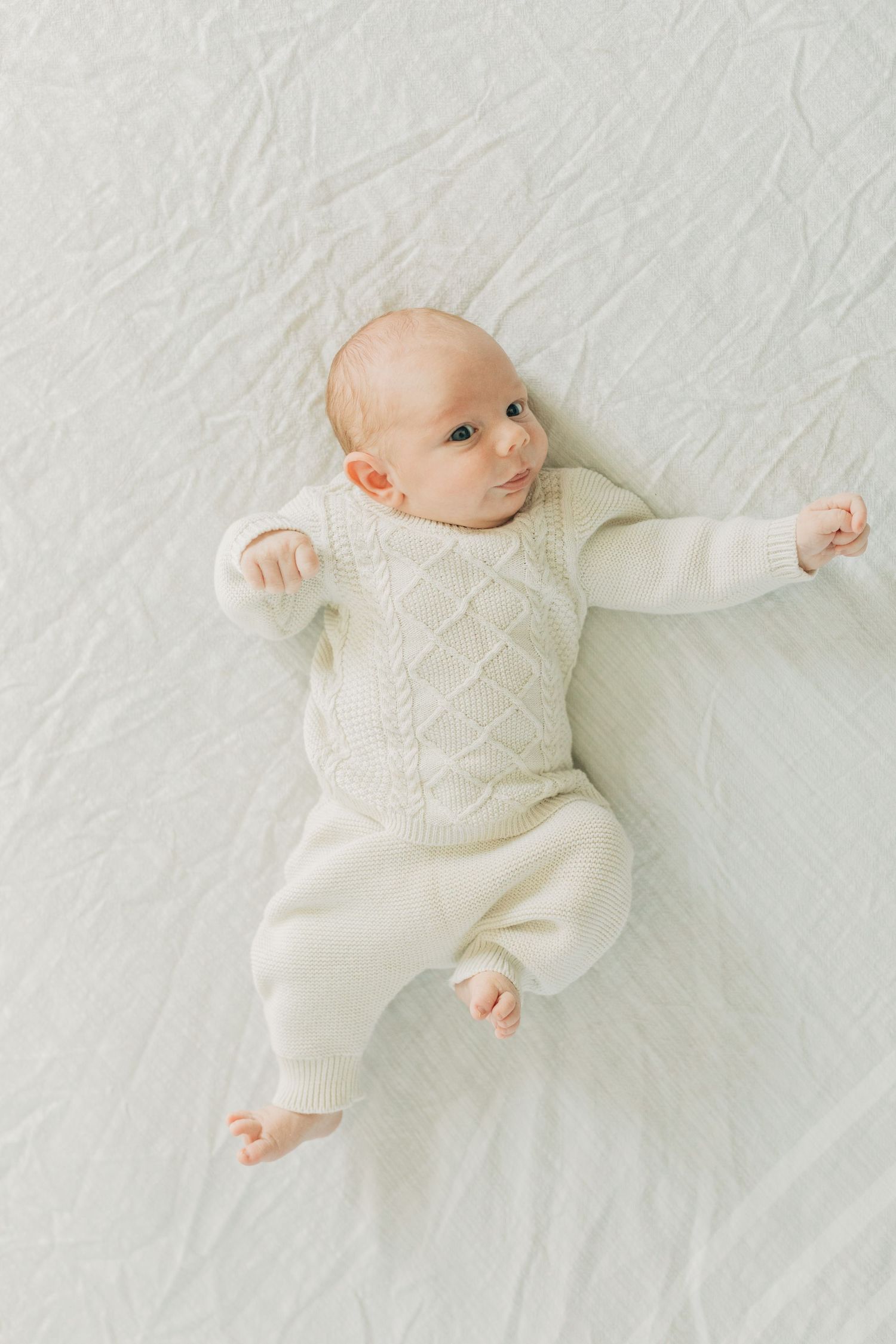 Baby in cozy white sweater stretches out on soft white surface.