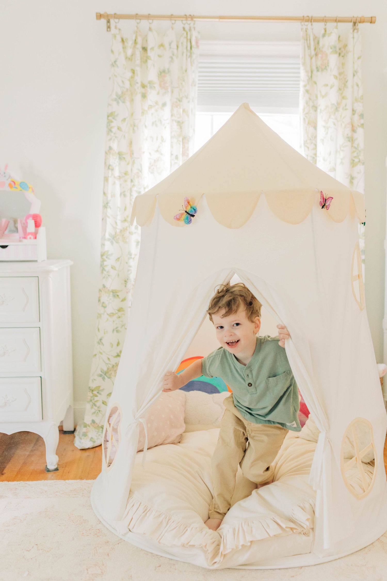 A cozy white teepee tent set up in a bright room with floral curtains and decorative butterflies.
