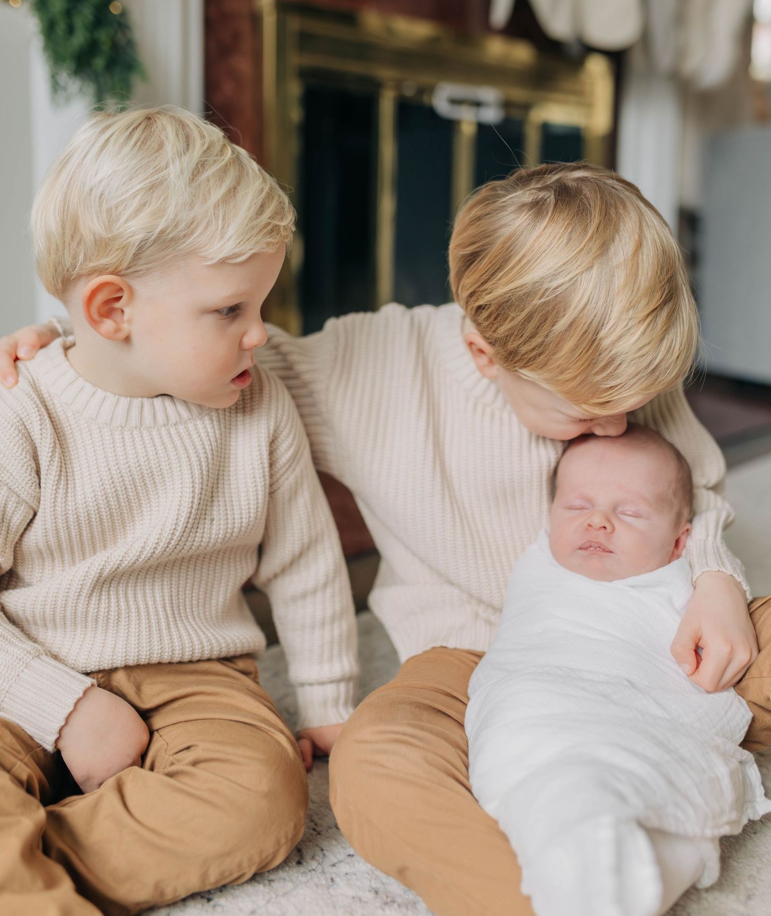 Two children in beige sweaters sit with a baby in white clothing near a decorated fireplace.