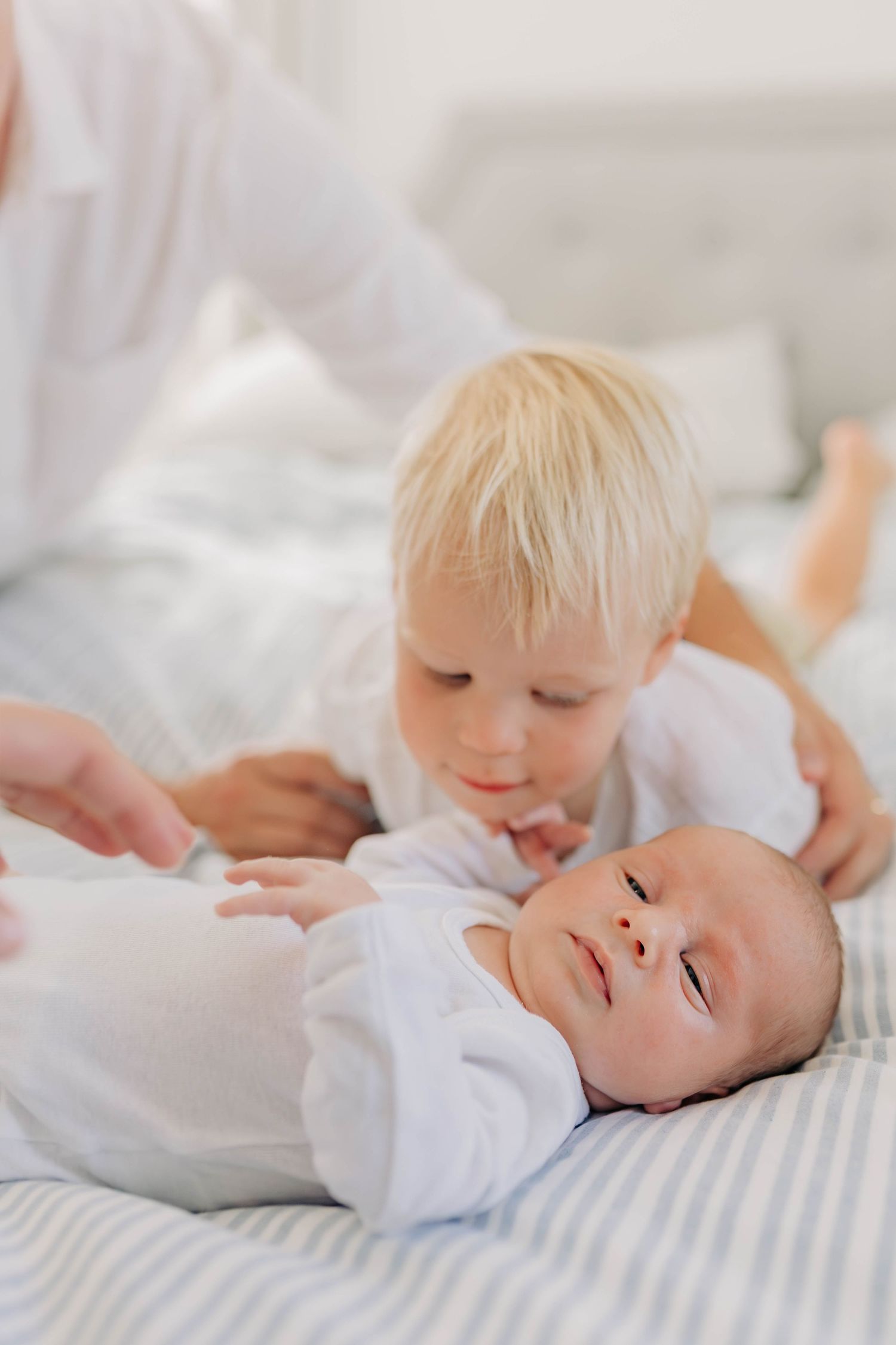 Two siblings share a sweet moment on white bedding as the older one looks at the baby.