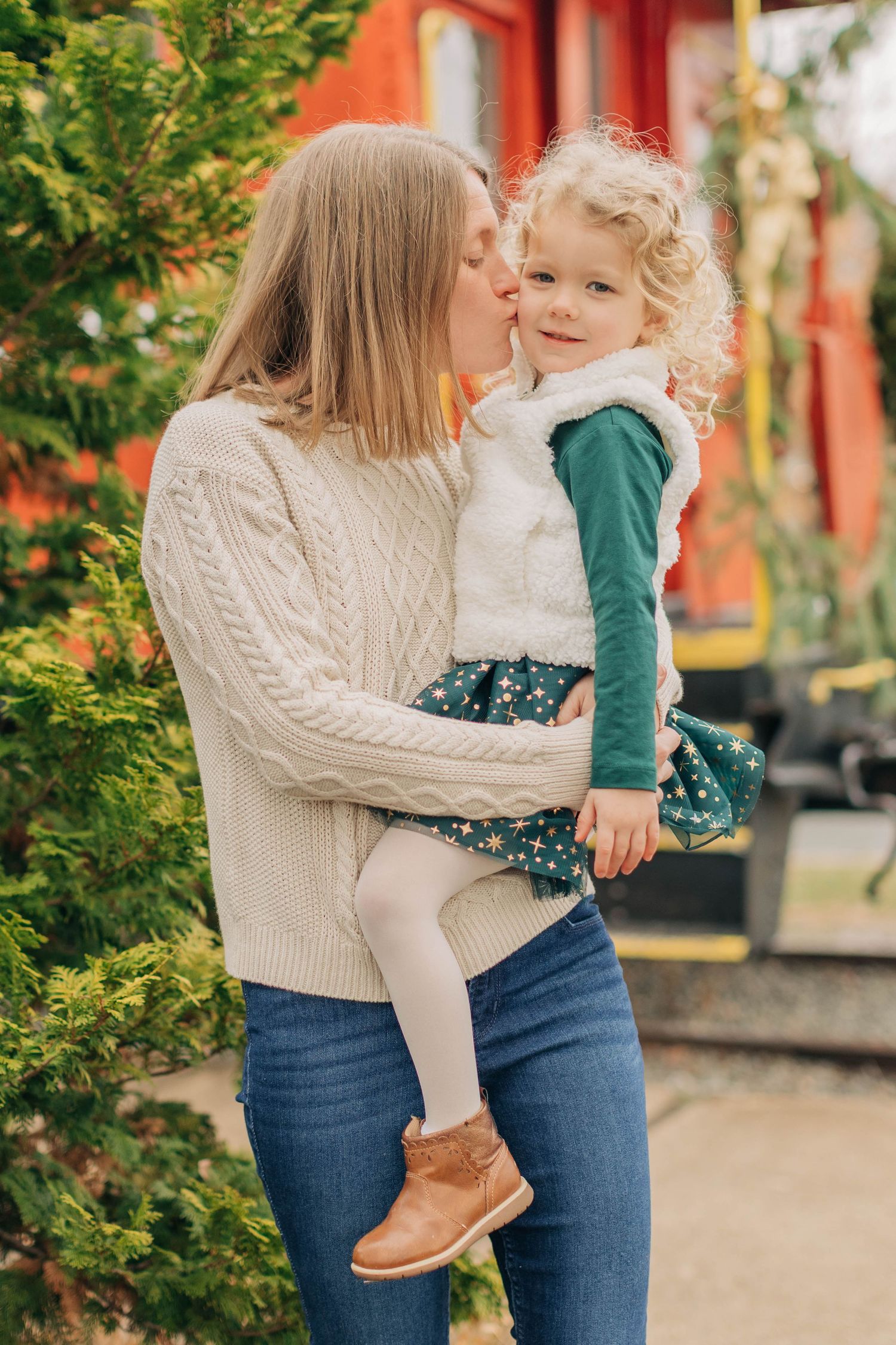 A mother in a cream sweater and jeans kisses her curly-haired toddler girl outdoors near green shrubs.