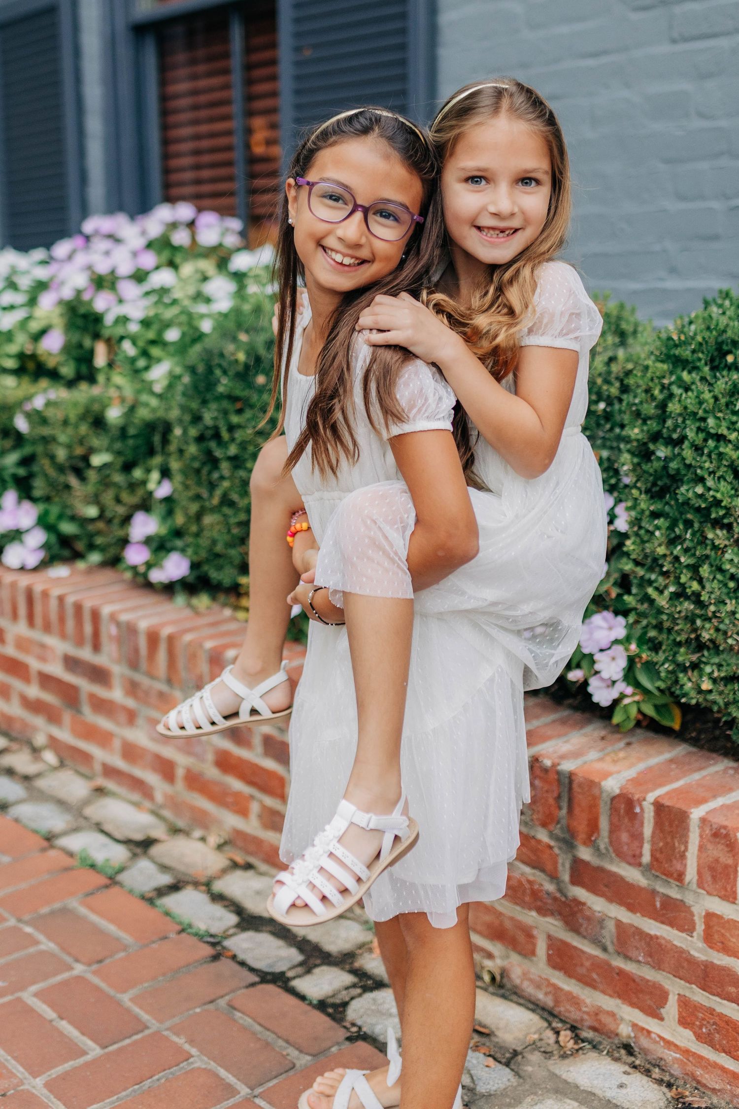Two young girls in white dresses pose outdoors, one giving the other a piggyback ride near a brick wall with flowers.