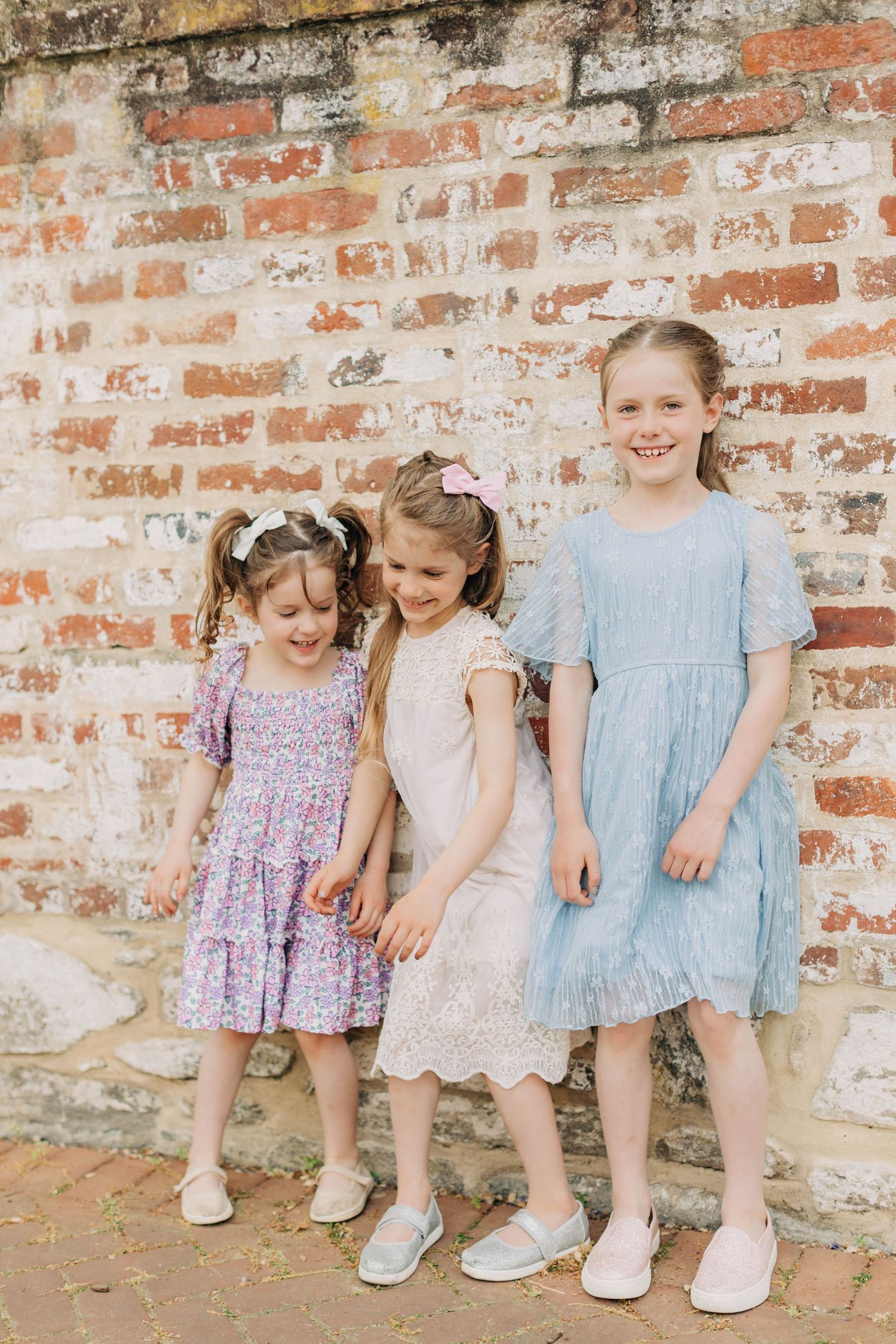 Three young girls in pastel dresses posing together against a rustic brick wall outdoors.