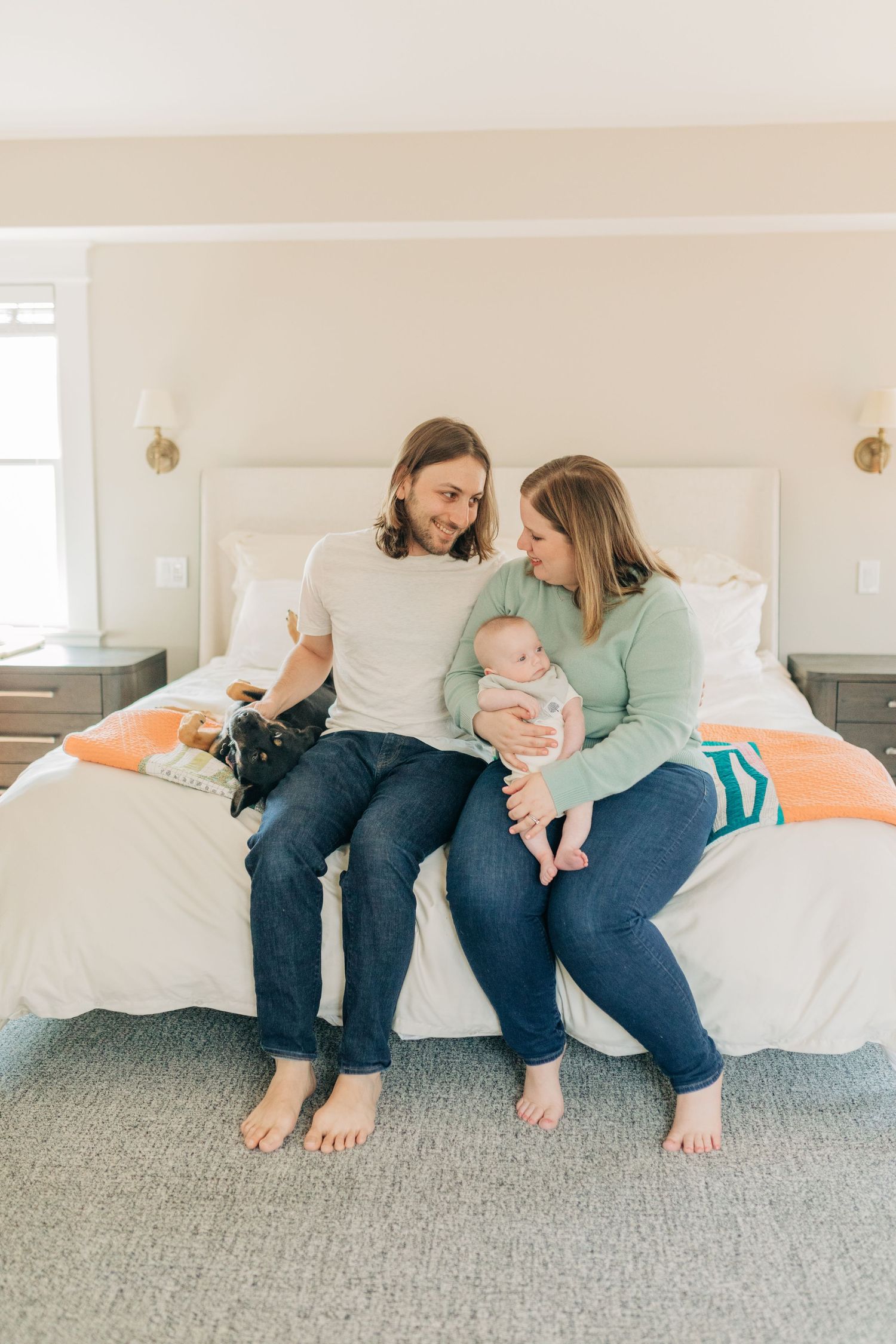 Family relaxes together on white bed with grey carpet in bright modern bedroom.
