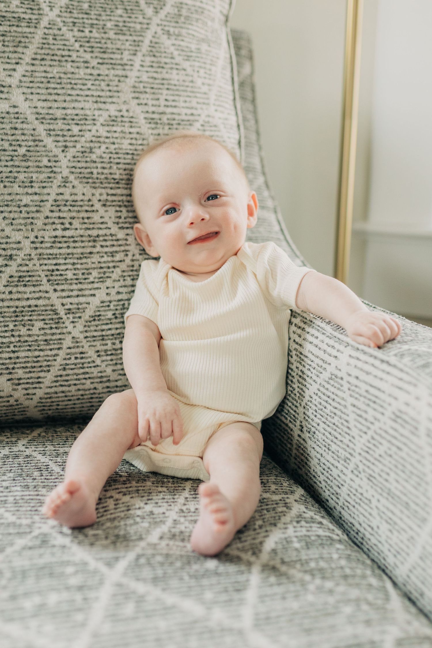 Baby in cream colored outfit sits on gray textured couch.