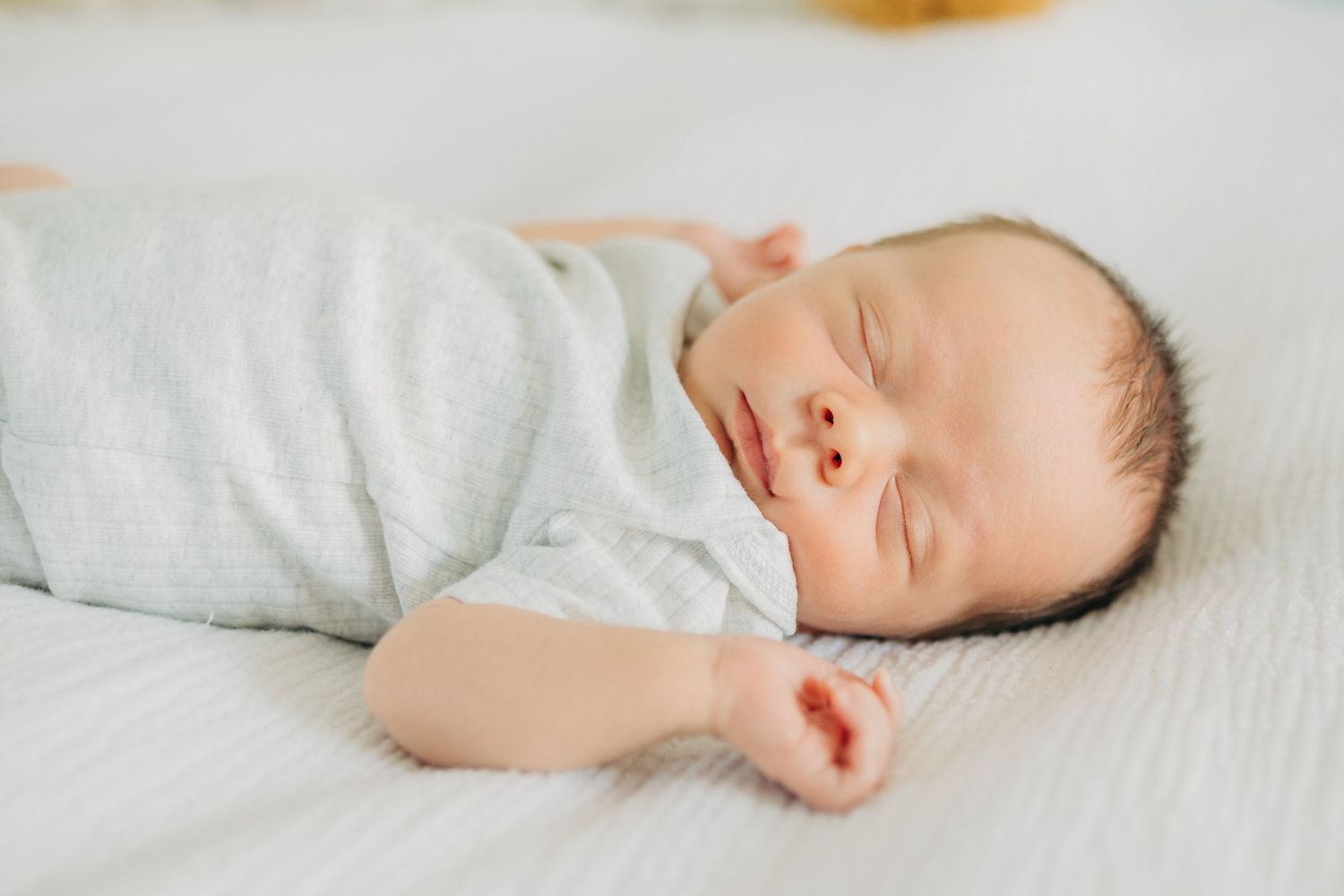 Sleeping newborn in light grey onesie on white bedding with natural lighting.