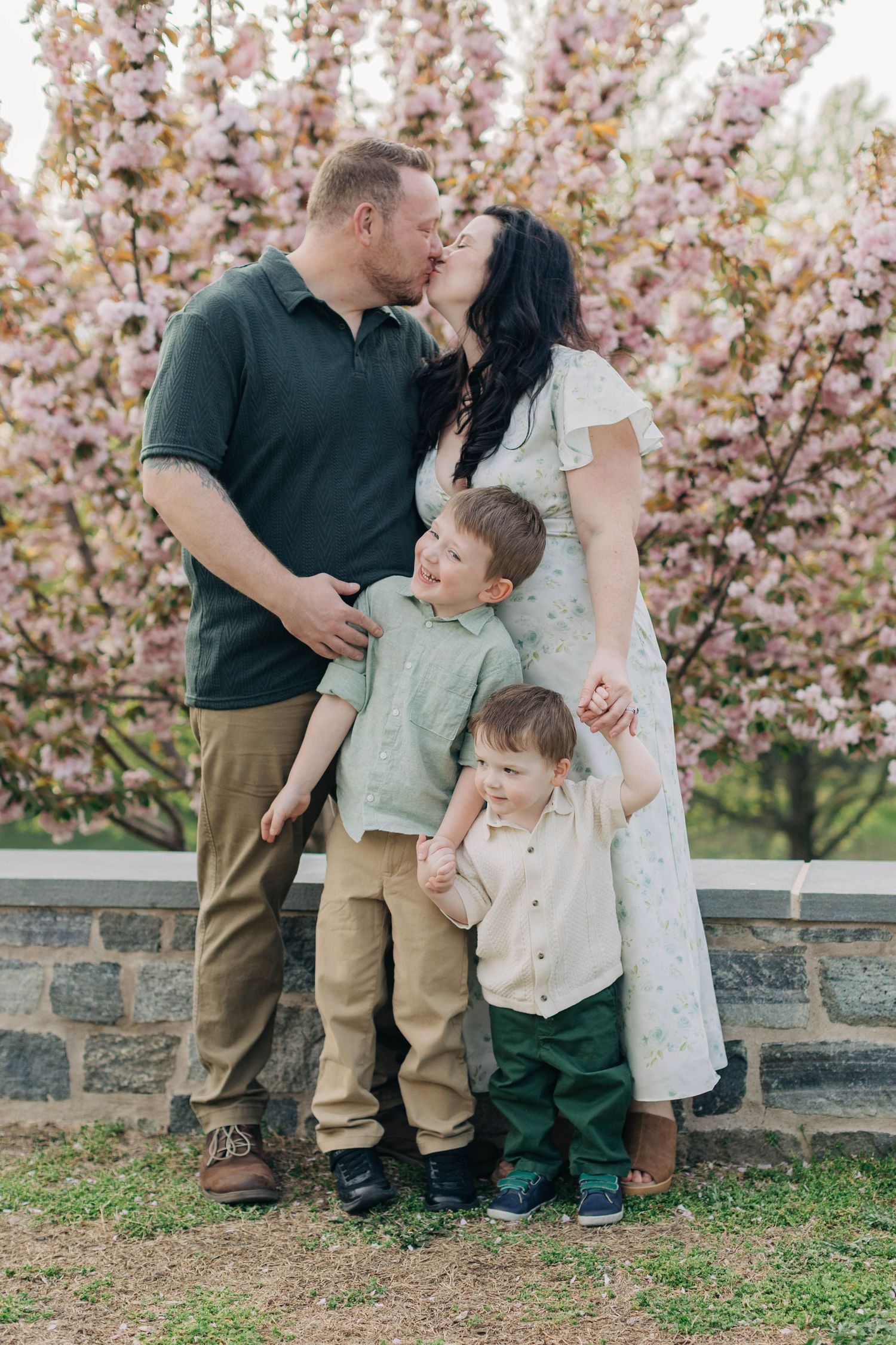 A family of four poses in front of pink cherry blossoms, parents kissing while two young boys stand between them.