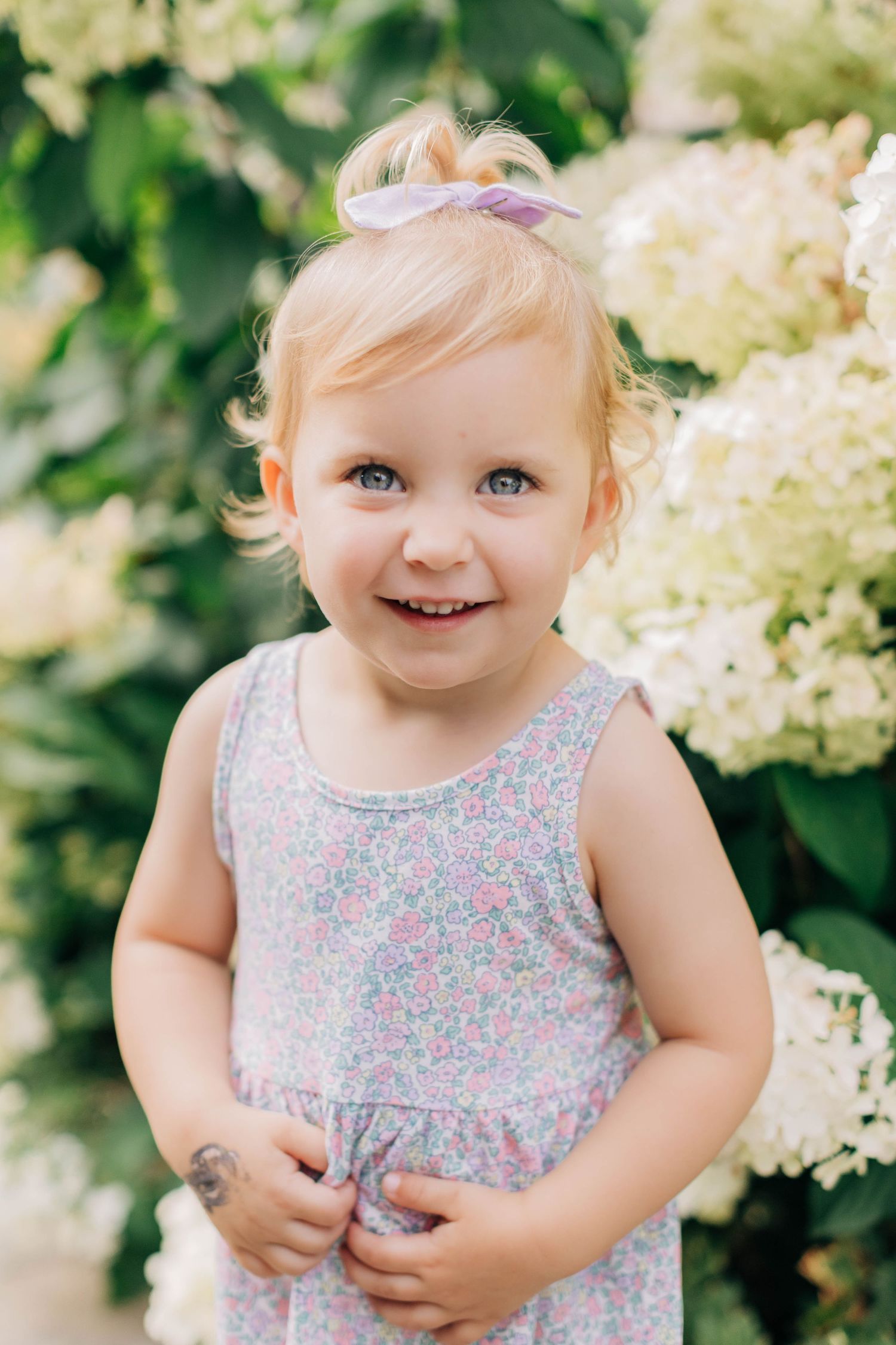 Smiling toddler girl with blonde hair and blue eyes wearing a floral dress, standing among white hydrangea flowers.