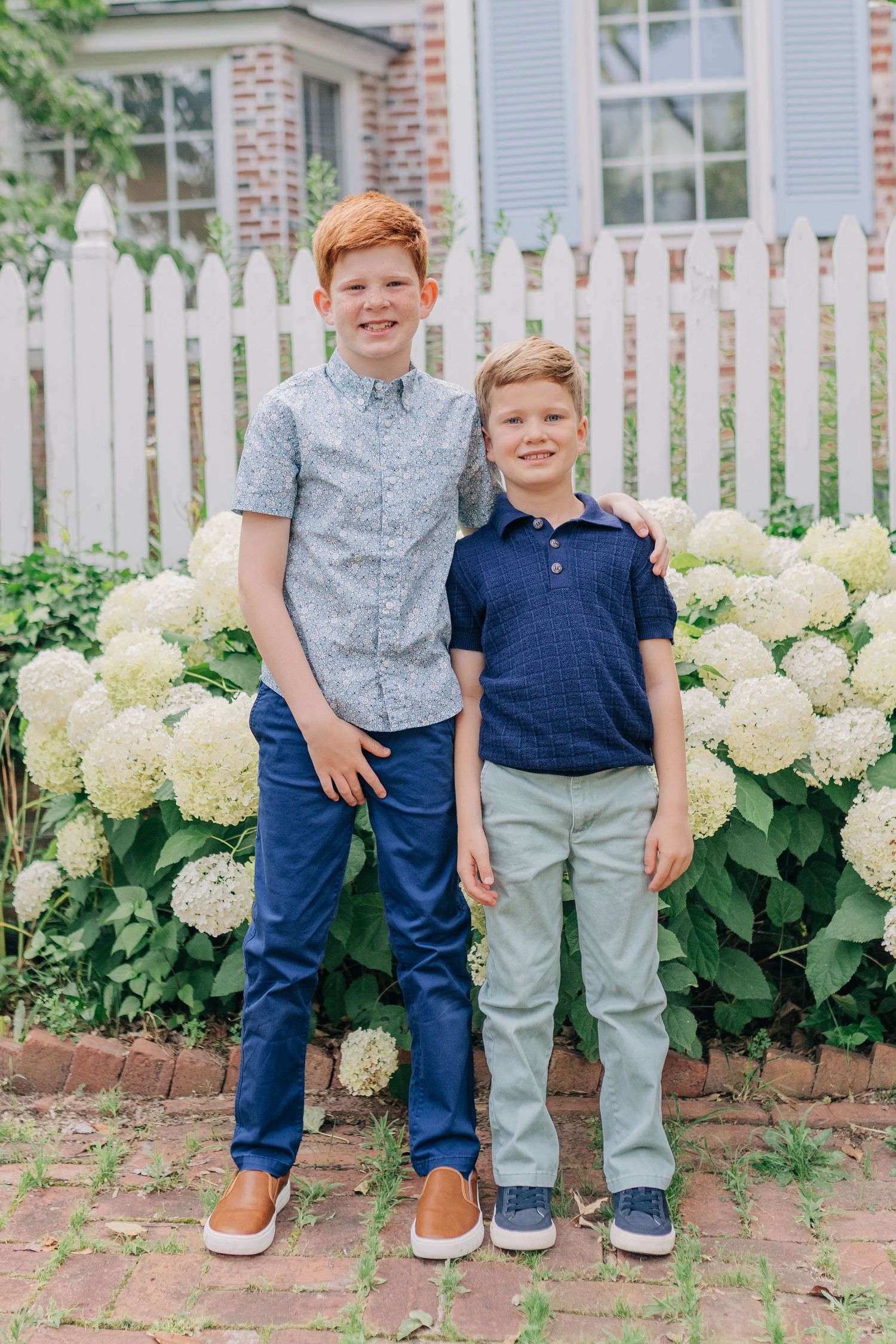Two smiling boys in navy outfits pose together in front of white hydrangeas and a picket fence.