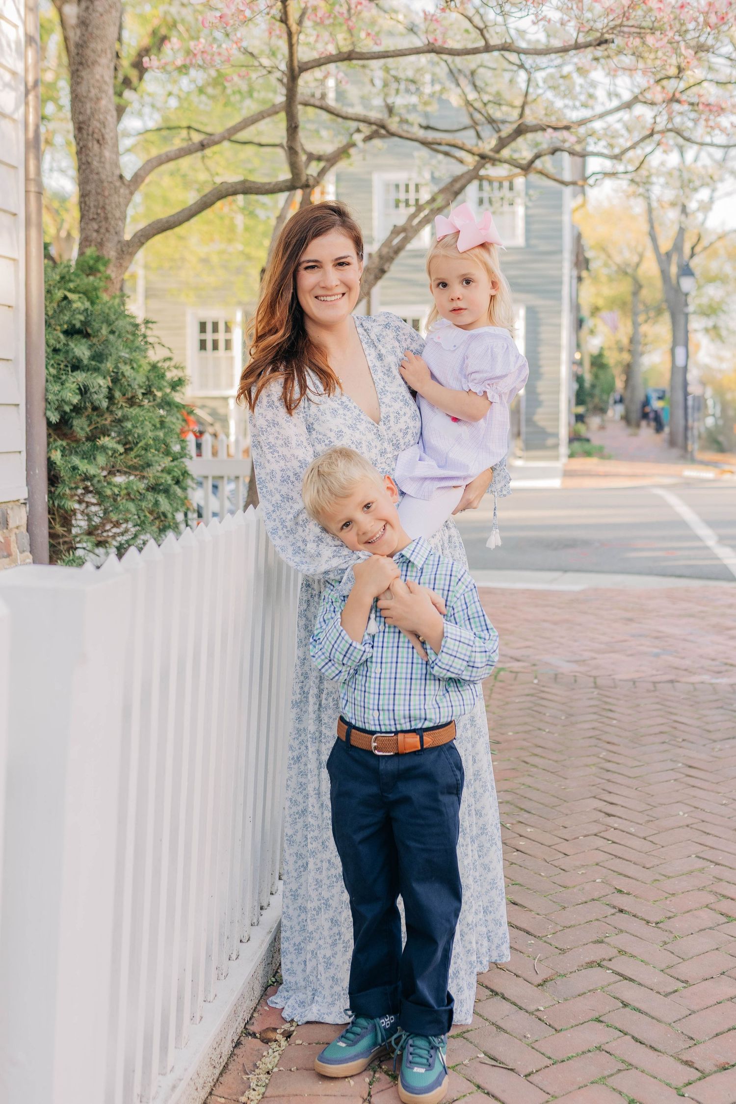 A smiling mother in a floral dress holds her toddler daughter while her young son stands beside her outdoors.