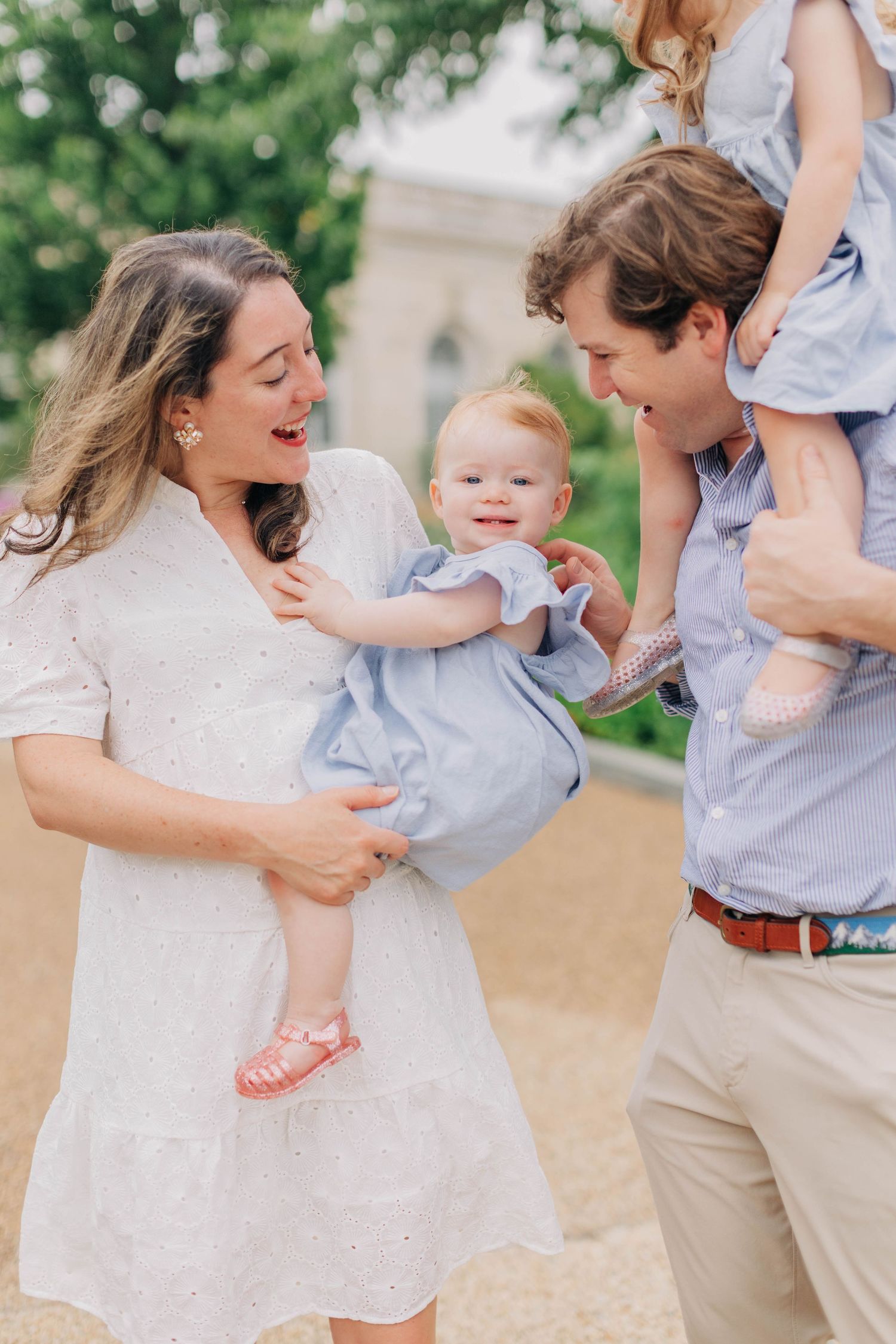 A happy family with two young daughters in matching blue dresses pose outdoors near a historic building.
