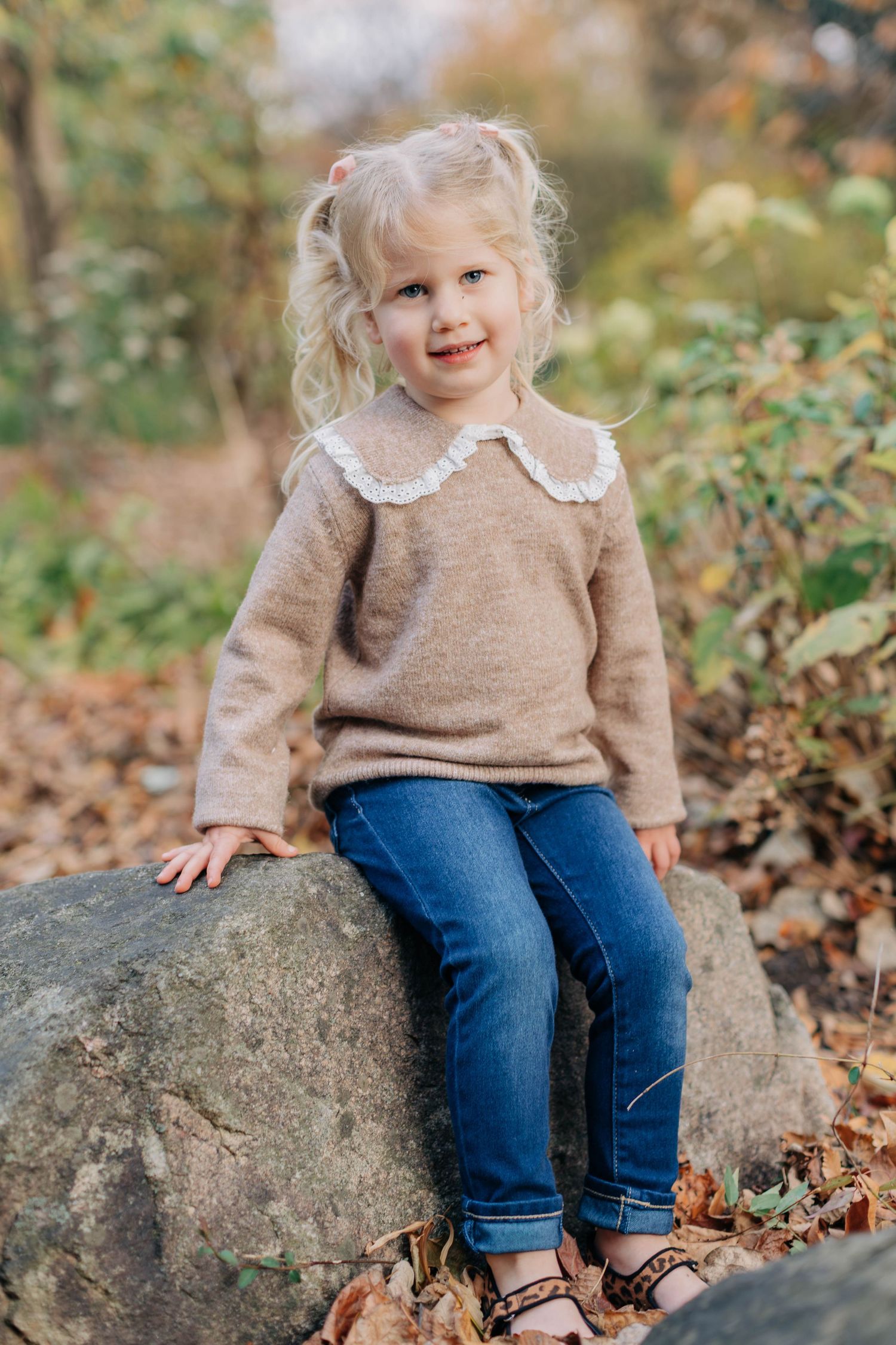 Smiling blonde toddler girl sitting on a rock outdoors in a beige sweater and blue jeans during autumn.
