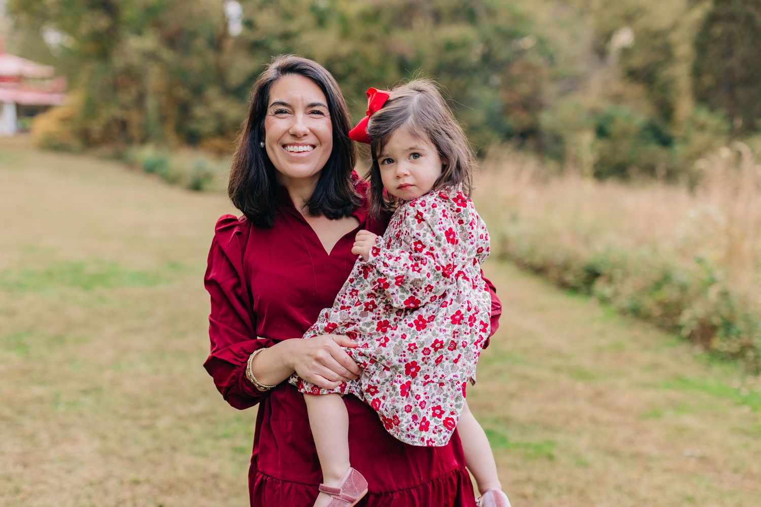 A smiling woman in a red dress holds a young girl in a floral outfit with a red bow outdoors.