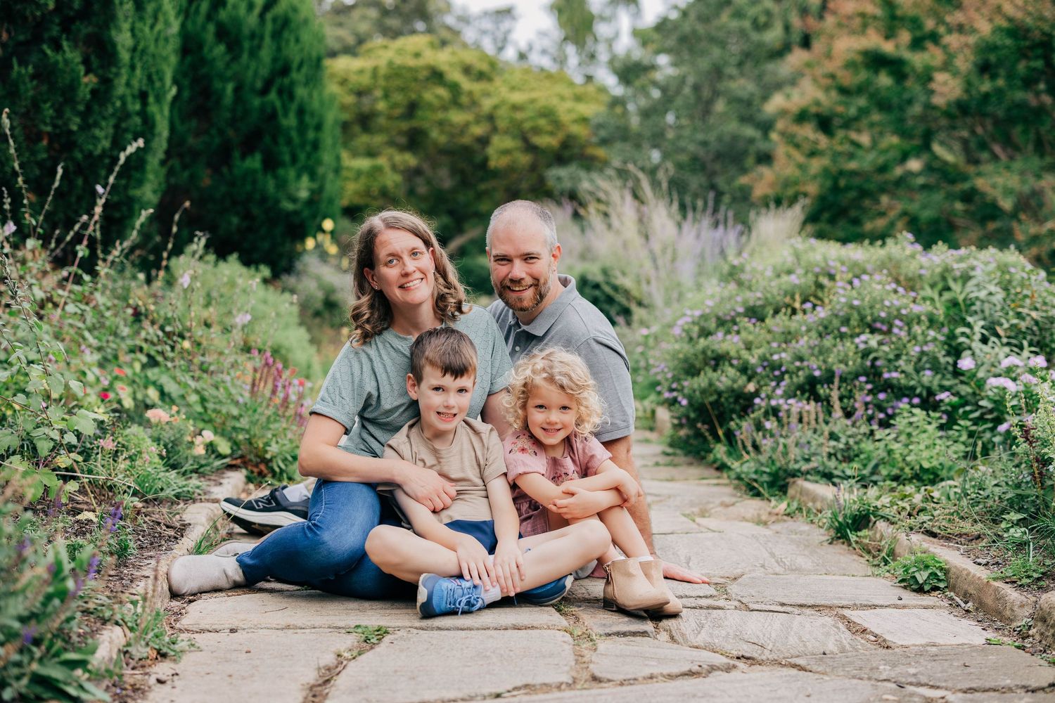 Smiling family of four, two parents and two young children, sitting together on a stone garden path surrounded by green plants and flowers.