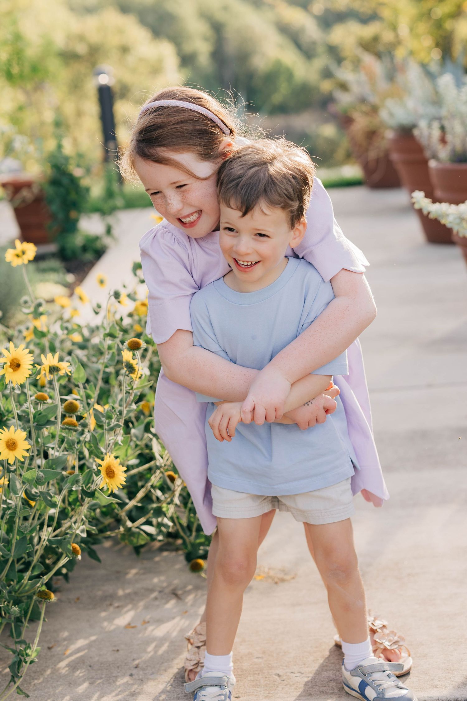 Two happy children hugging outdoors near yellow flowers, girl in purple dress embracing smiling boy in blue shirt.