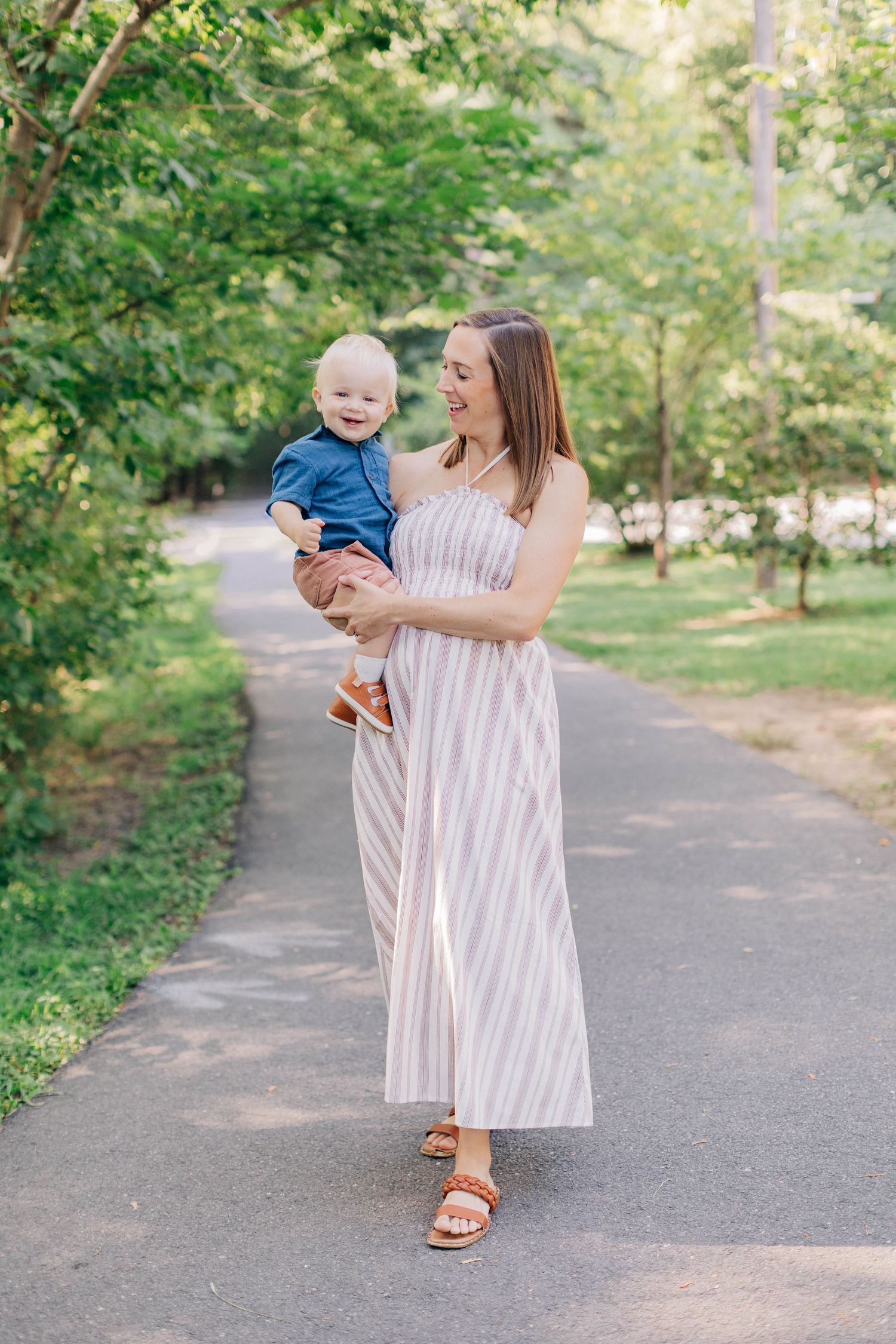 A smiling mother in a striped maxi dress holds her happy baby boy while walking on a tree-lined path.