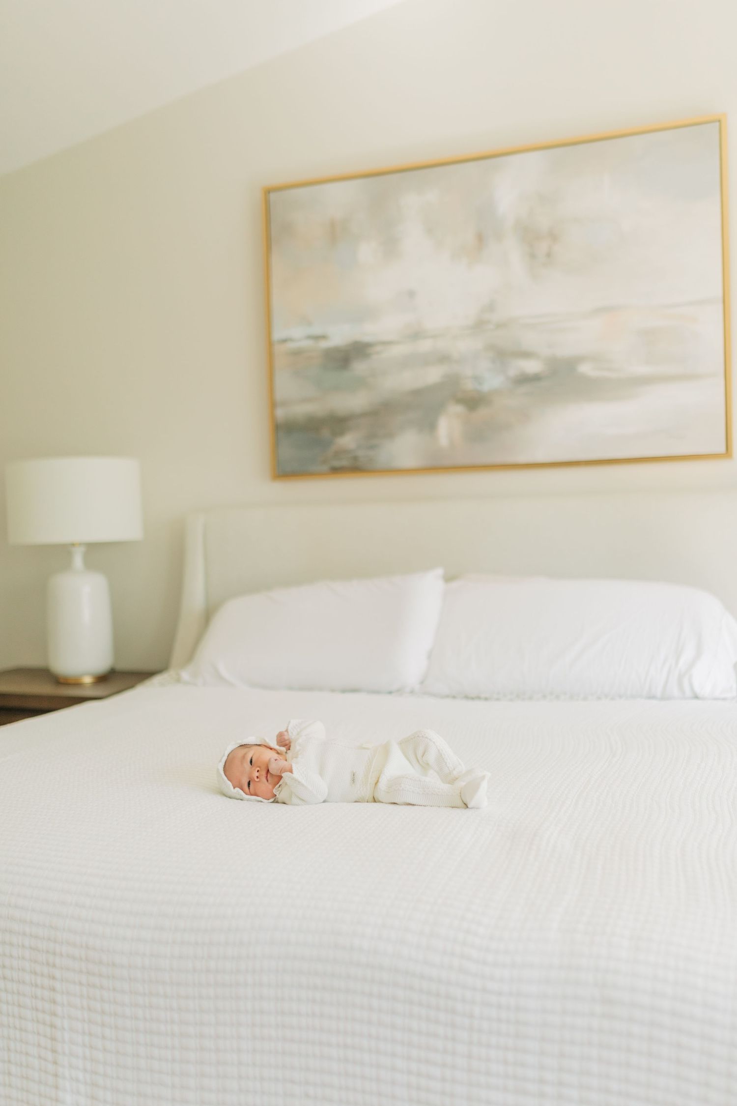 Infant resting on white bed with abstract artwork and table lamp in background.