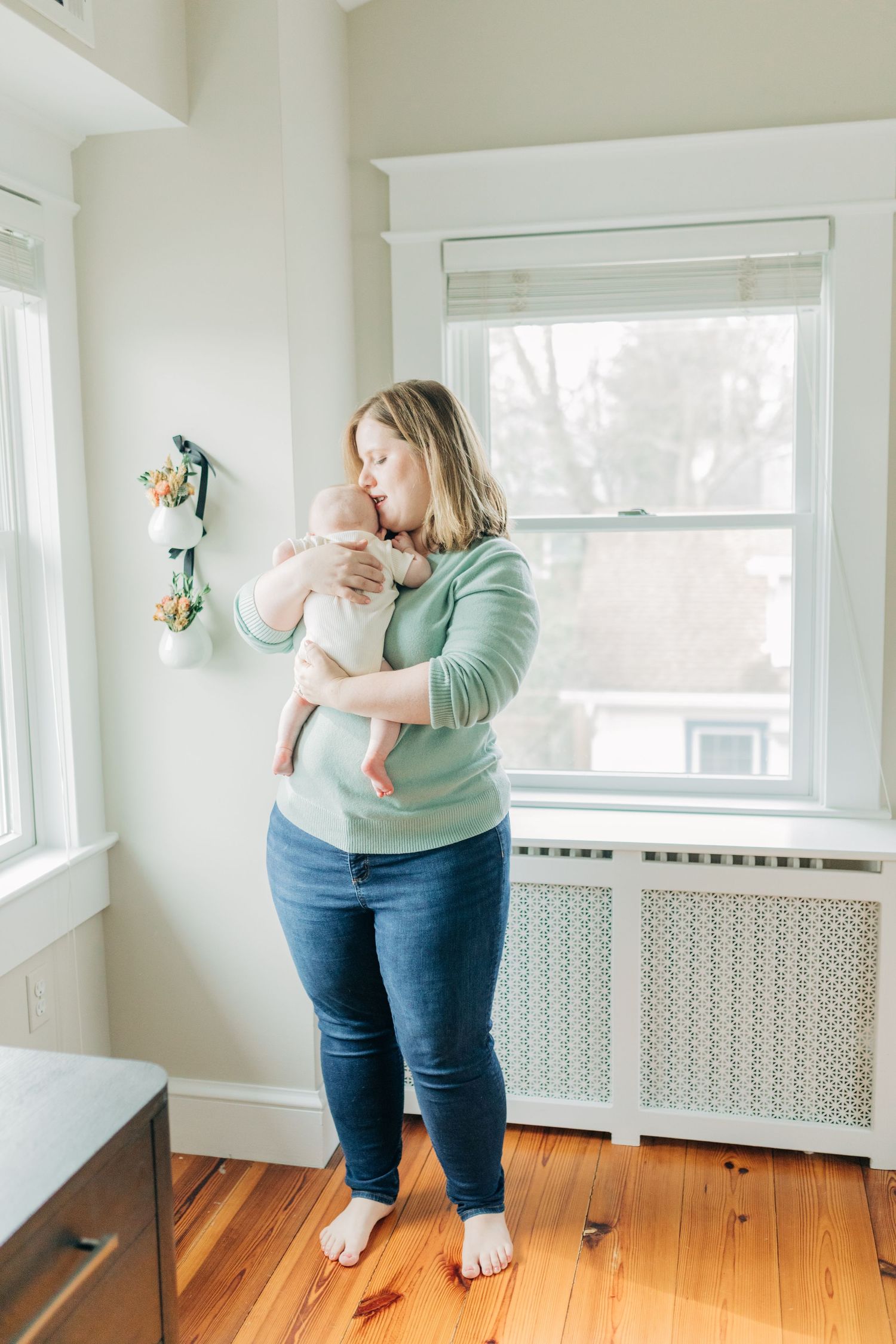 Parent in mint sweater holds baby in bright nursery with wooden floors.