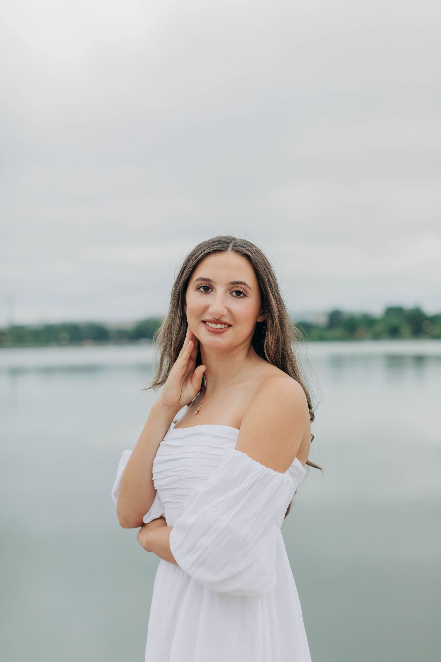Off-shoulder white dress portrait by reflecting pool with cloudy sky.