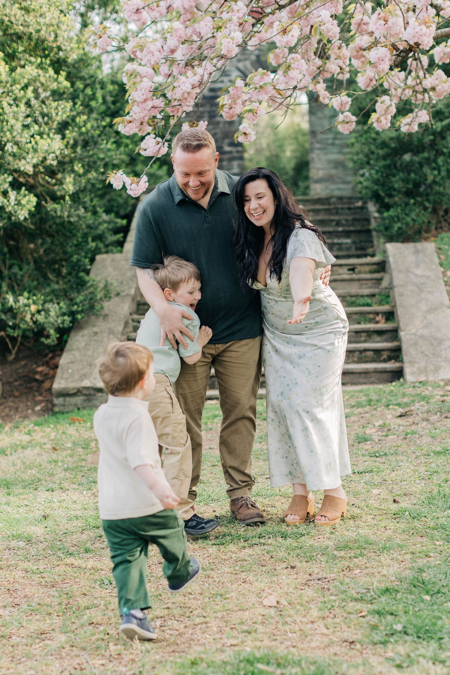A happy family plays together under pink cherry blossoms in a lush garden, with parents laughing as children run to them.