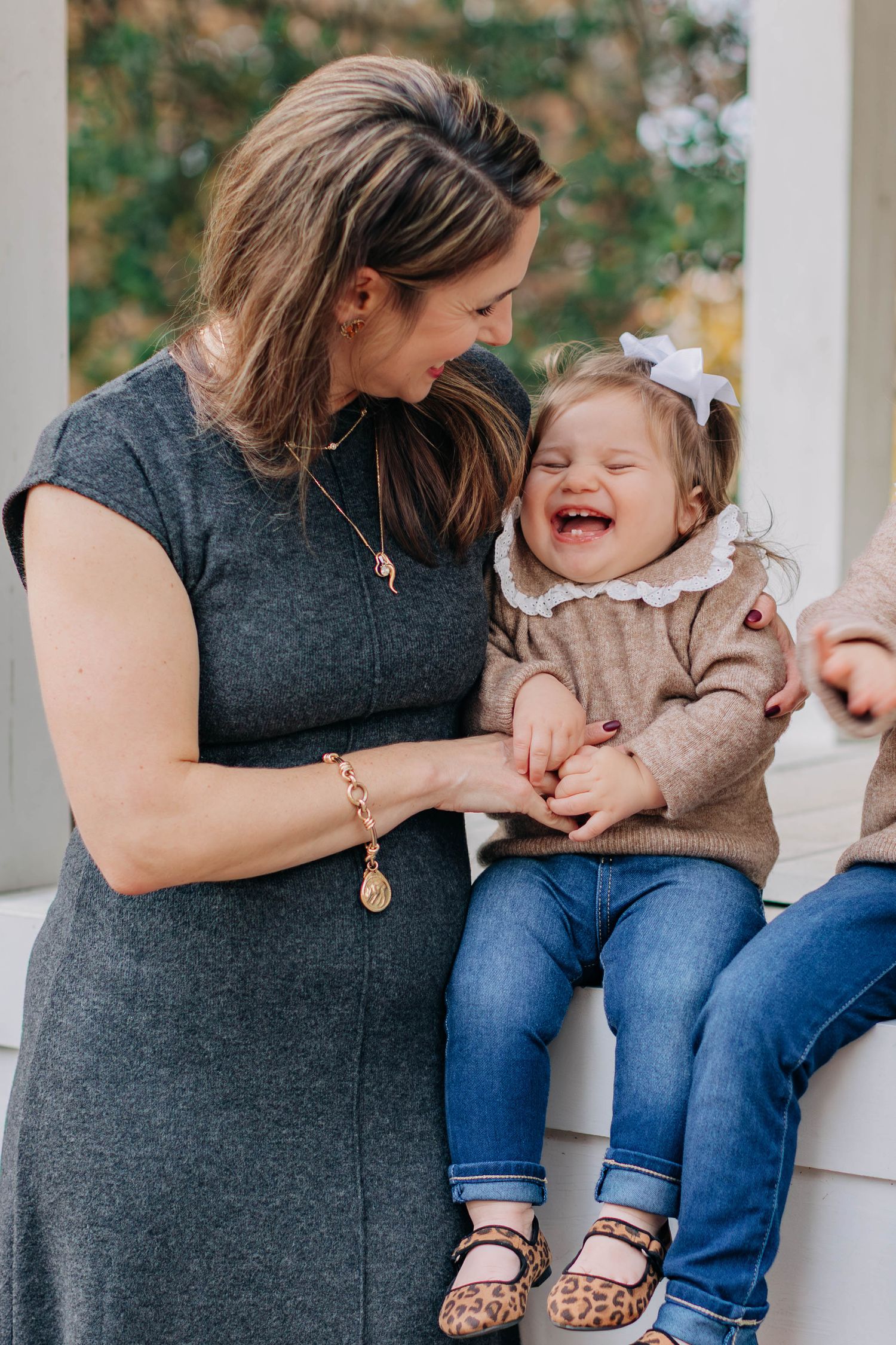 A woman in a gray dress laughs with a toddler girl in a bow and leopard shoes sitting on a white ledge.
